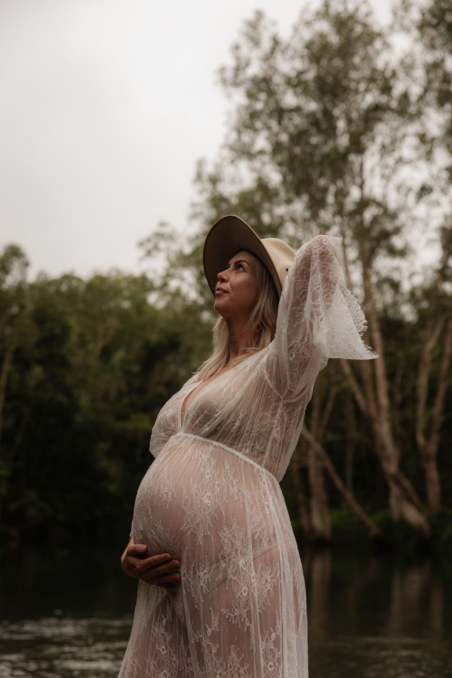 Maternity photo of a pregnant woman in a sheer lace dress standing by a lake at dusk.