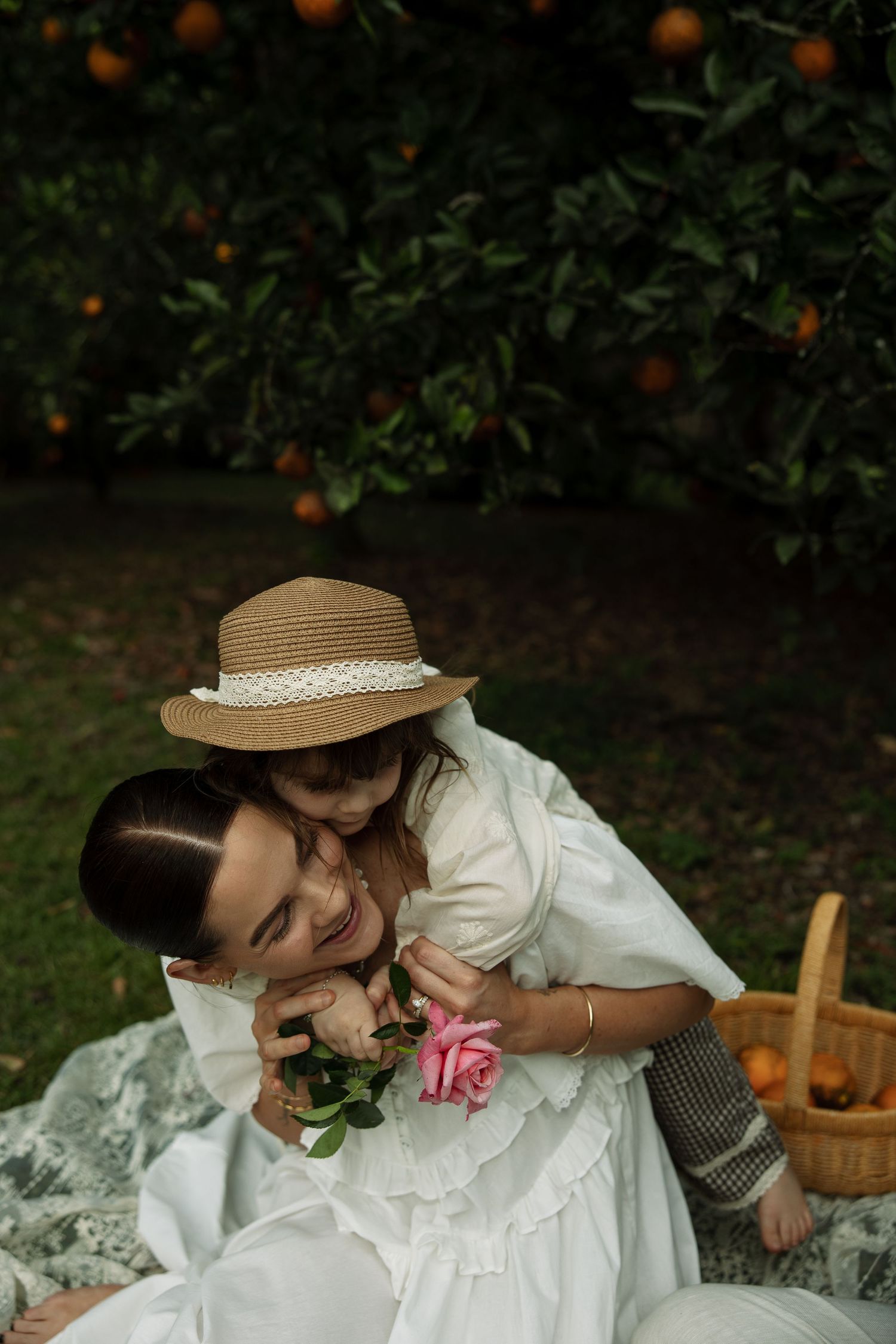 Sweet candid moment in a garden featuring white dress and straw hat with orange fruit trees in background.