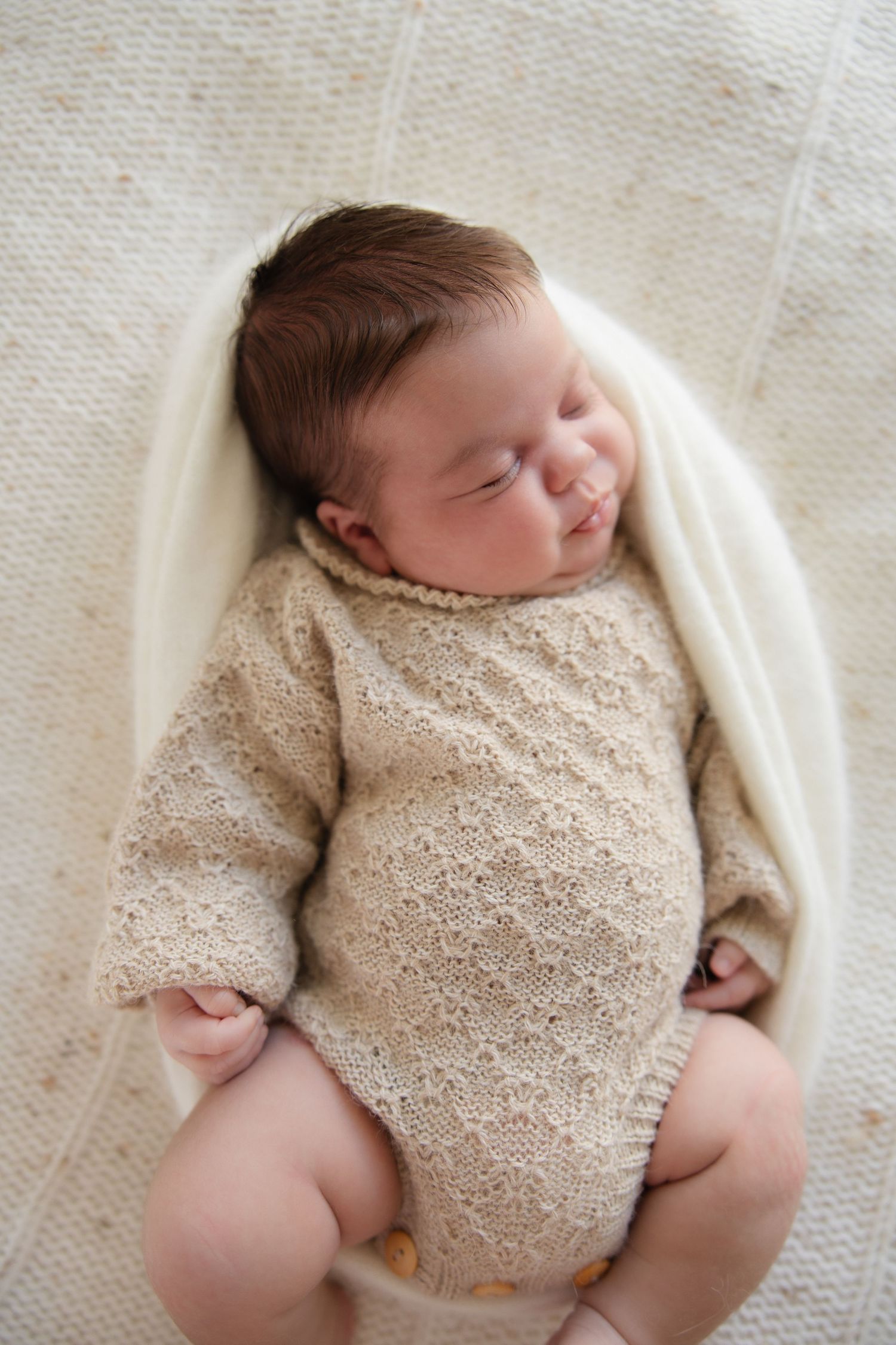 A newborn baby in a textured beige outfit smiles while sleeping on white fabric.