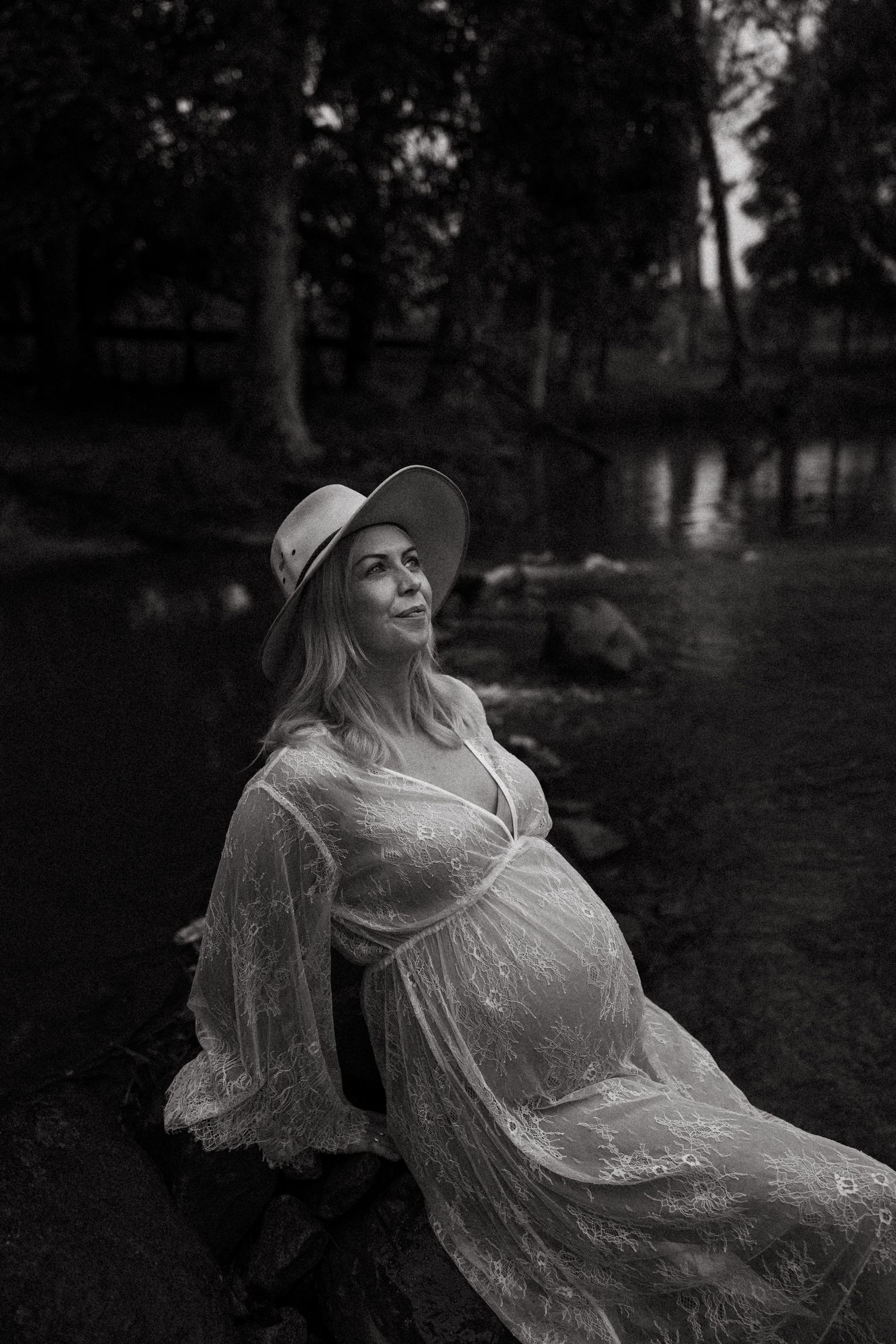 Dreamy black and white maternity photo of an expectant mother in a lace dress resting by a pond in a wooded setting.