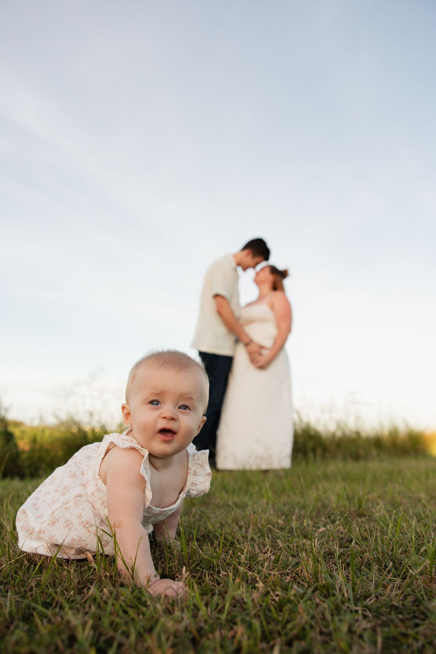 Family portrait in a grassy field with a baby crawling in foreground and couple embracing in background.