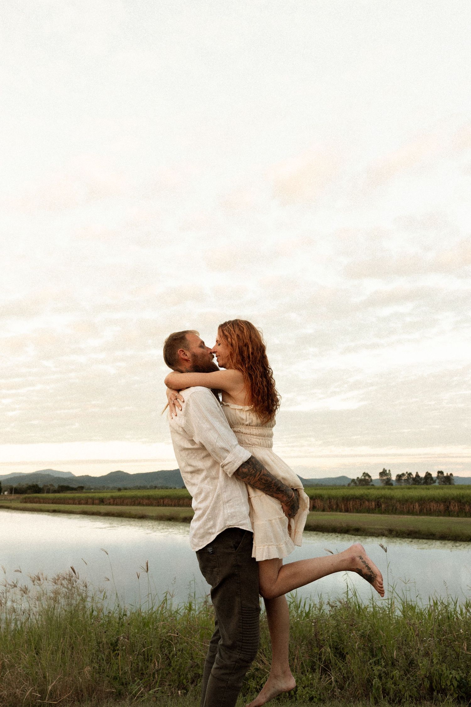 A romantic couple embraces by a serene lake at sunset with mountains in the distance.