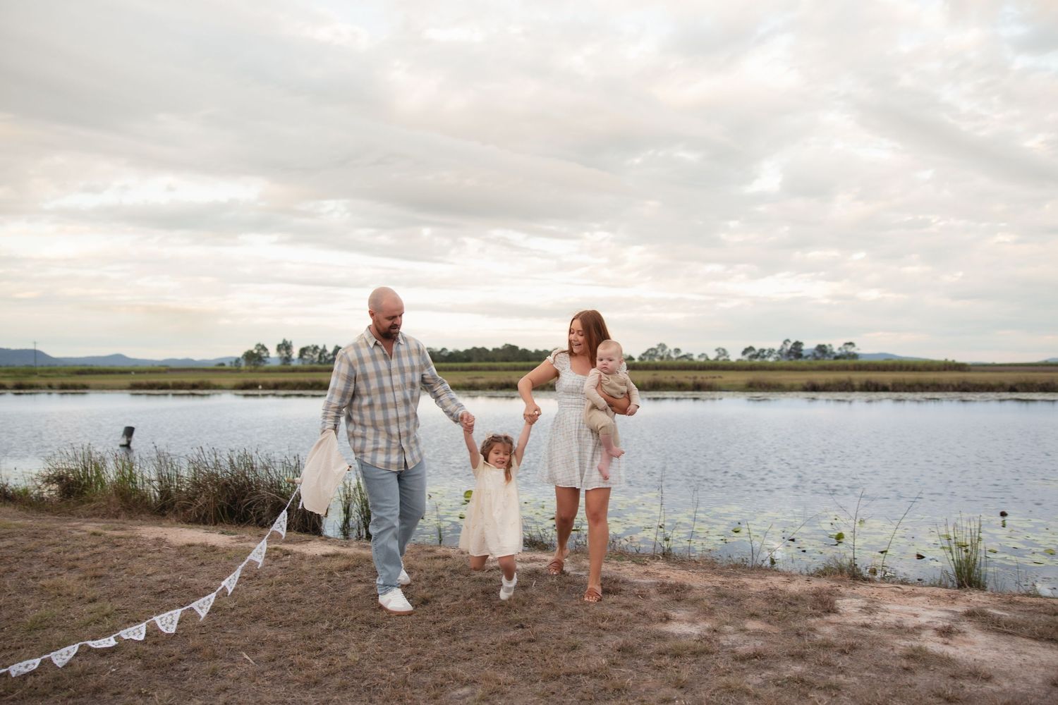 Family stands together holding hands at the edge of a tranquil lake during sunset with string lights in the foreground.