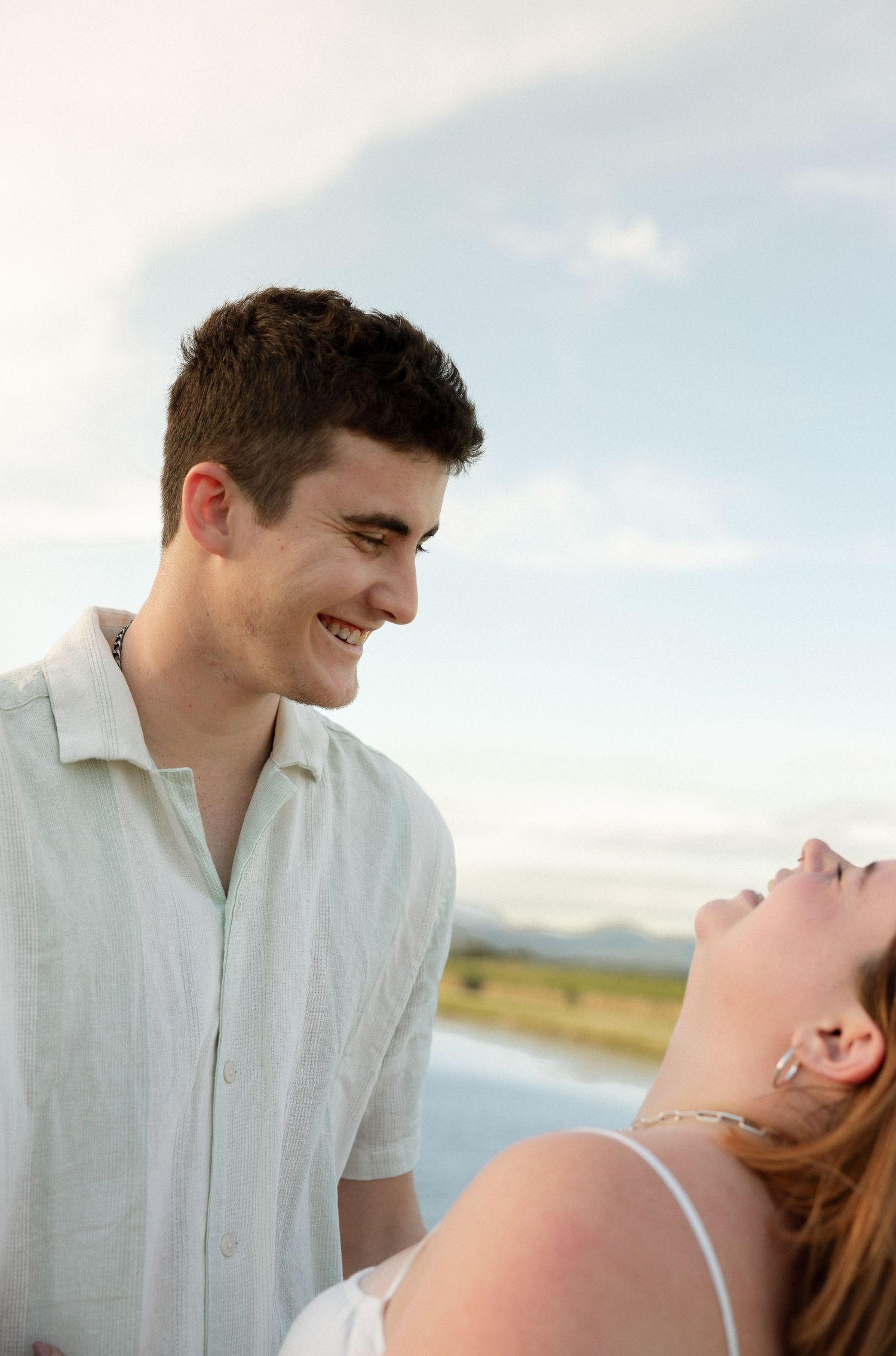 A couple shares a joyful moment outdoors with blue sky and mountains in the background.
