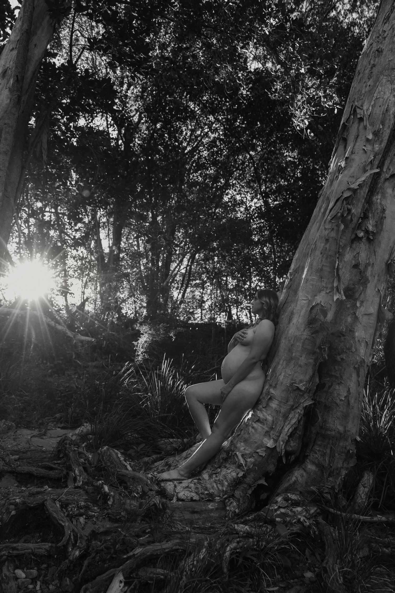 Artistic black and white nude photograph of person resting against a tree trunk with sunlight filtering through leaves.