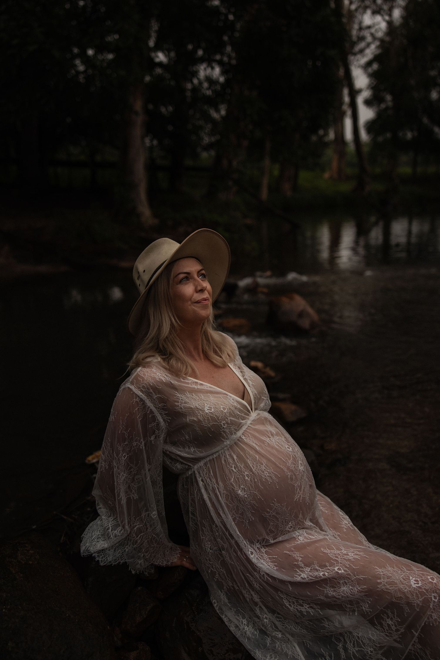 A pregnant figure in a sheer white dress and hat reclines by a pond in a moody forest setting at dusk.