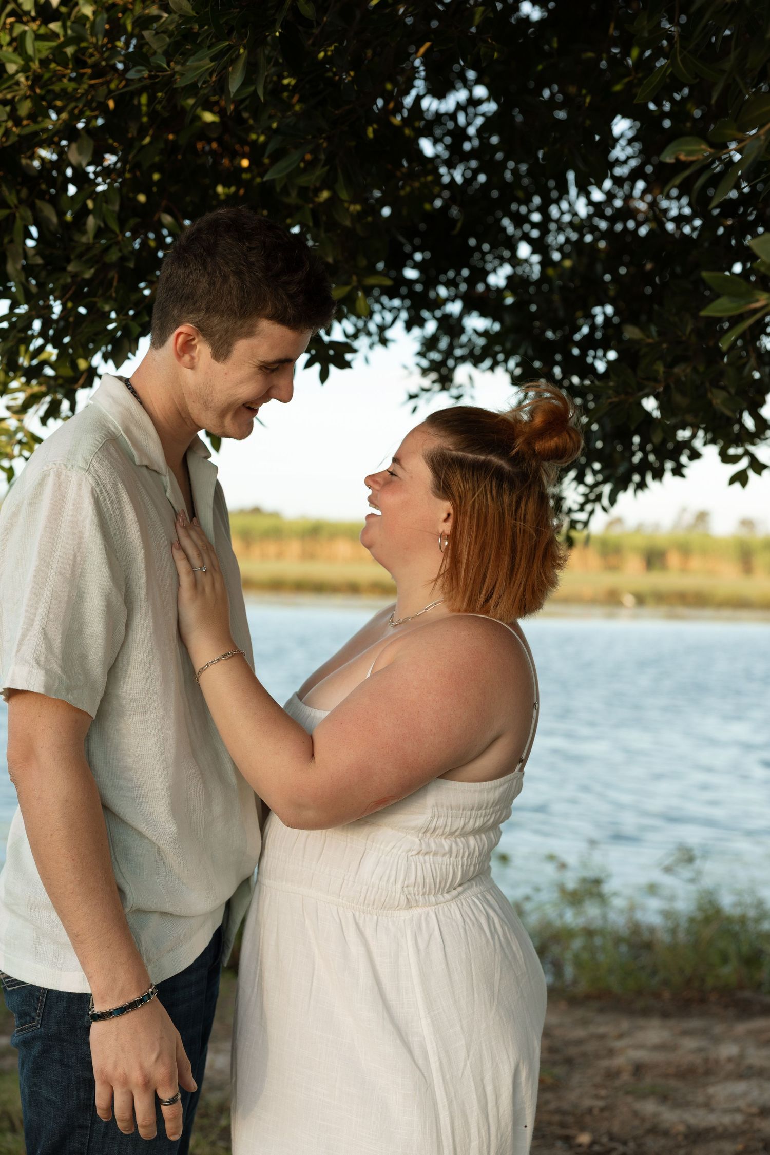 A romantic moment between a couple standing under a tree by a lake during golden hour.