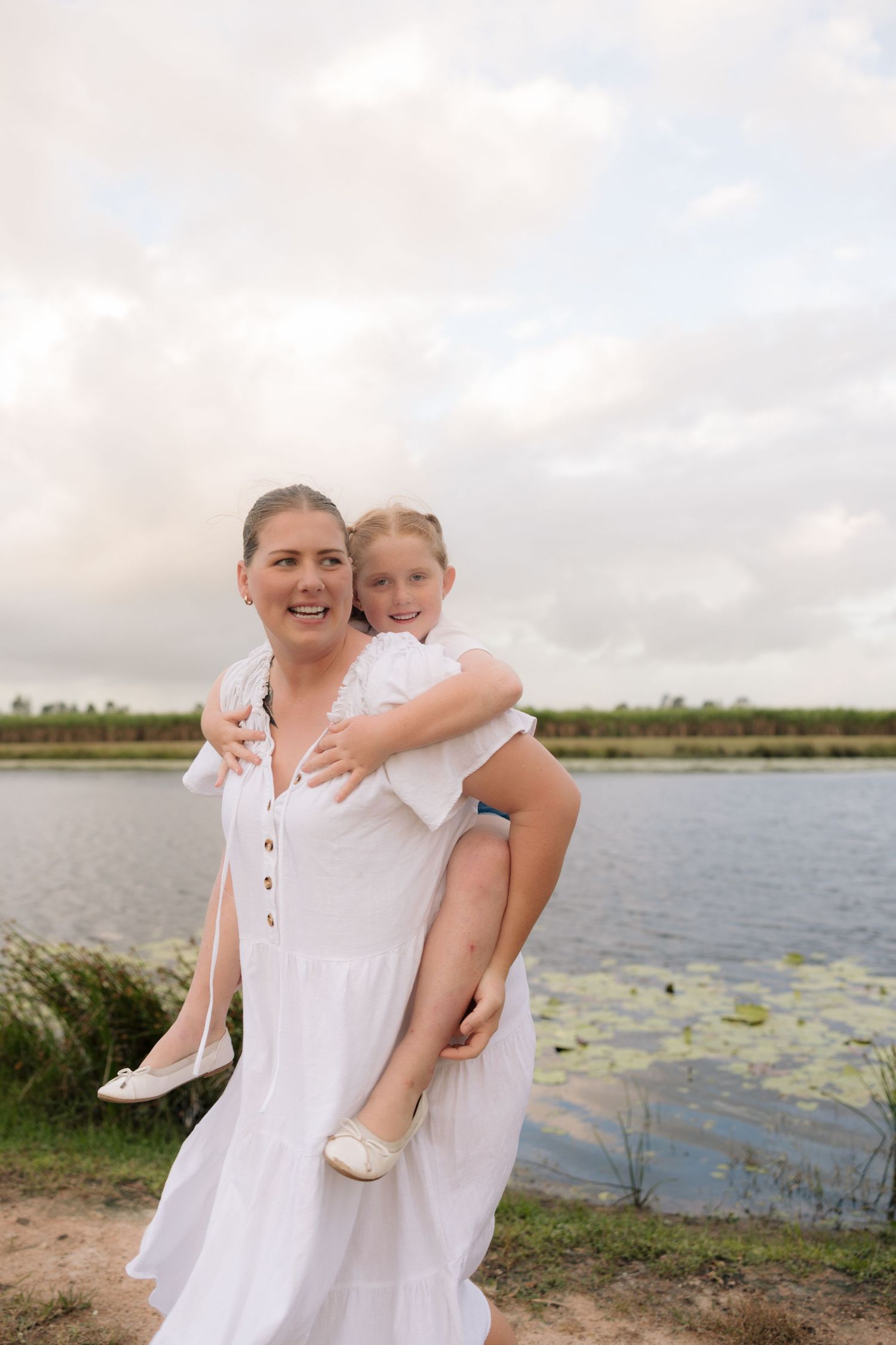 White-dressed figure carrying child on back near waterfront with cloudy sky.