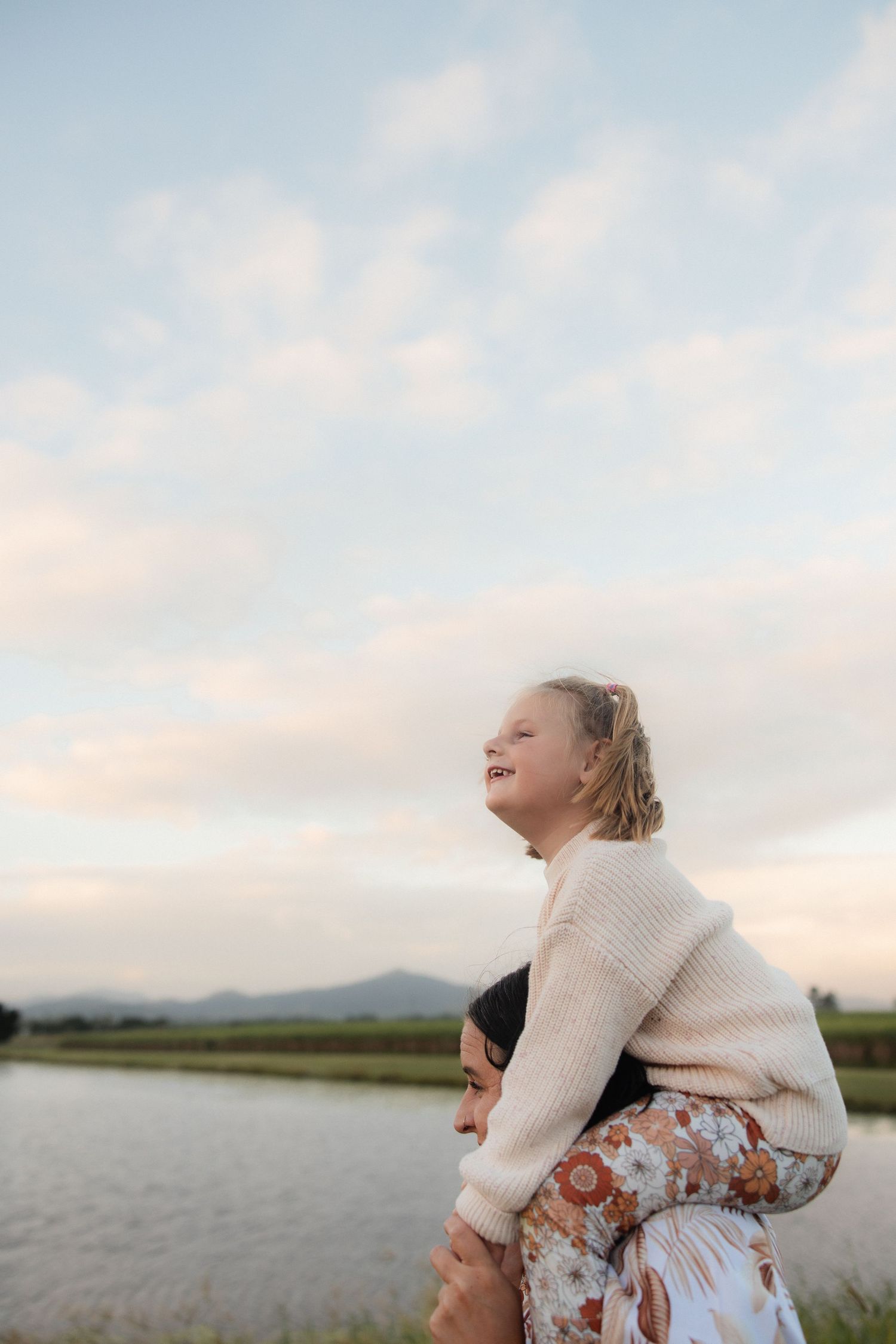 Child riding on shoulders by the lake during golden hour.