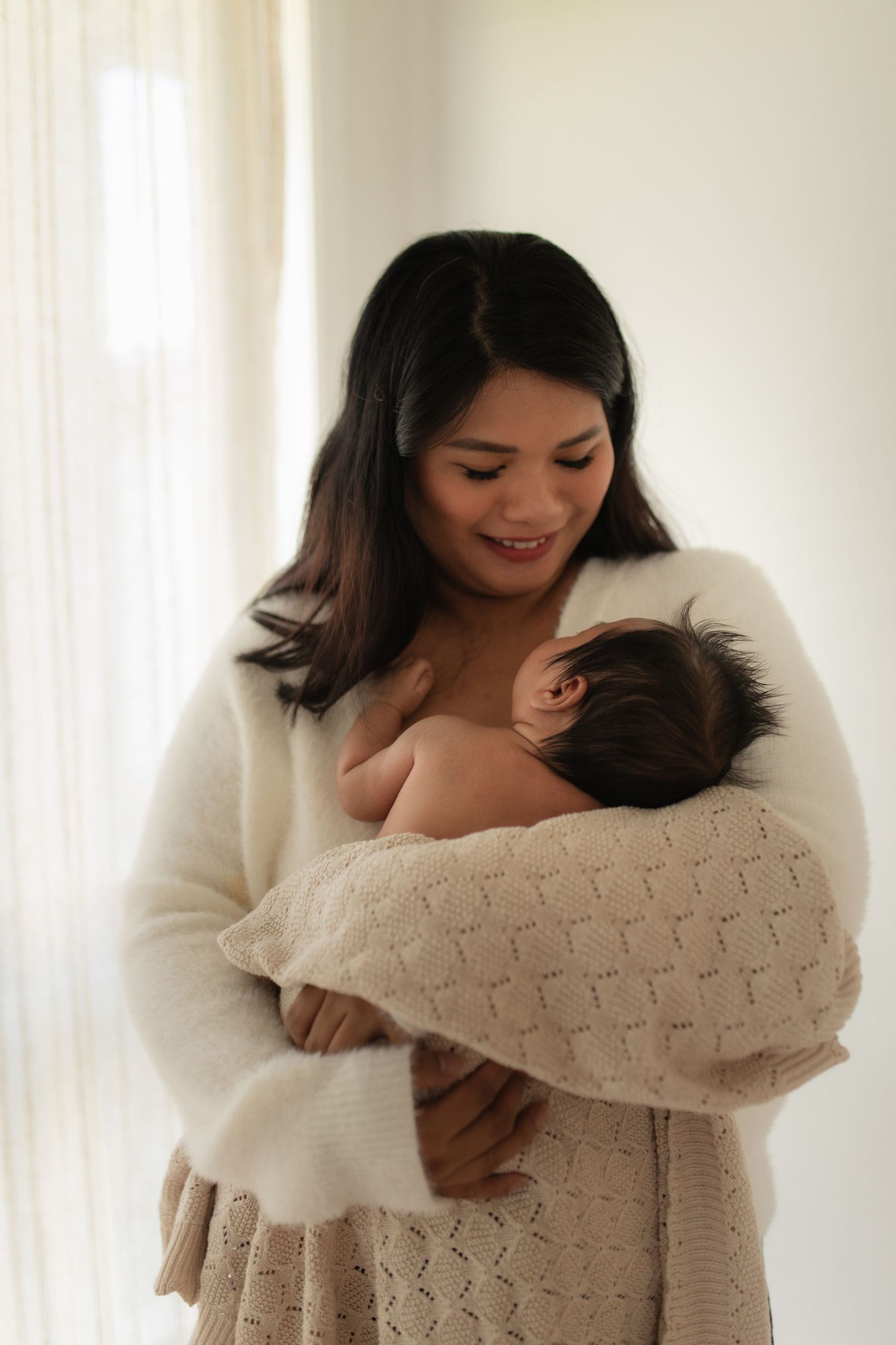 A mother in a white sweater tenderly holds her nursing baby wrapped in a knit blanket.