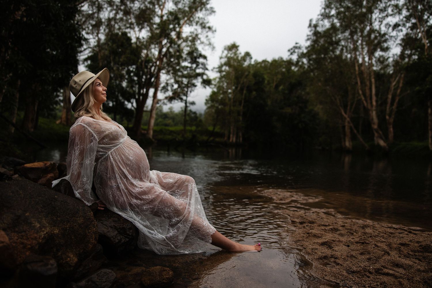 Person in elegant white lace dress sits at edge of dark river as sunlight filters through trees in moody forest setting.
