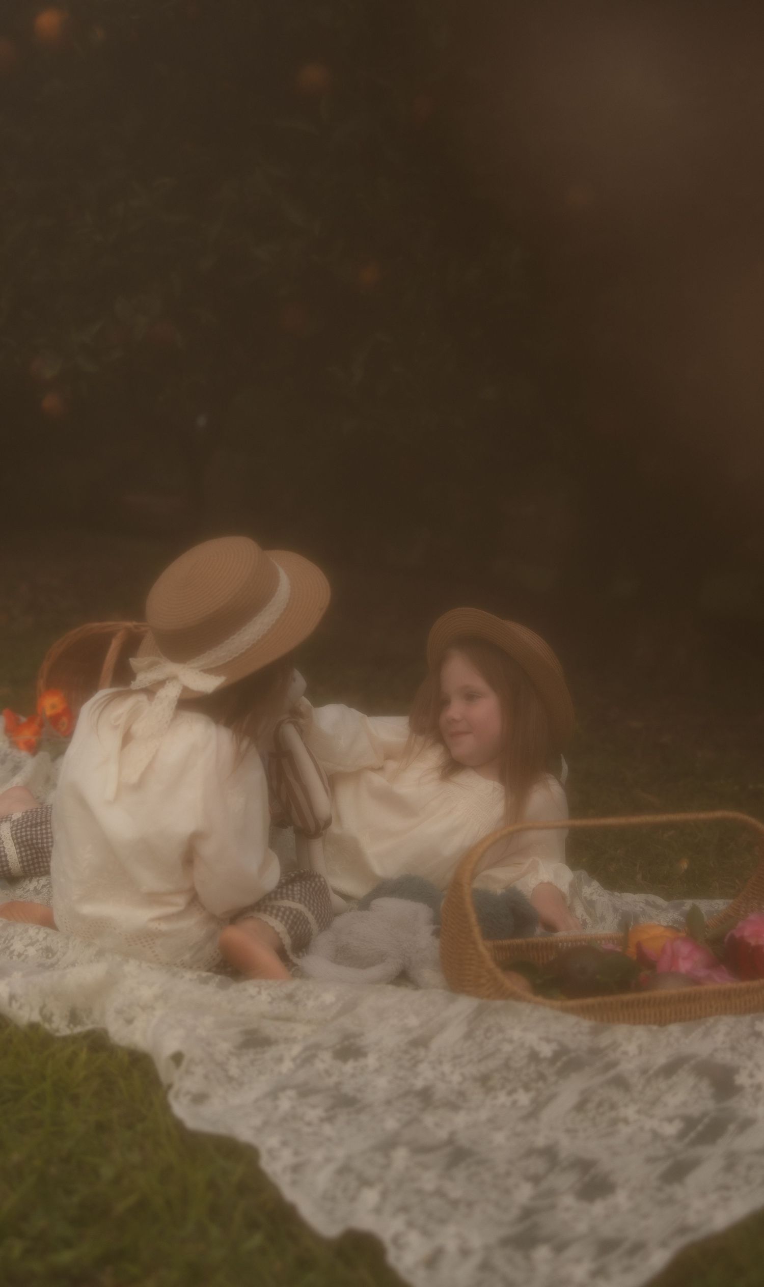 Two figures in vintage white dresses share an intimate picnic on a lace blanket in a dreamy, ethereal garden setting.