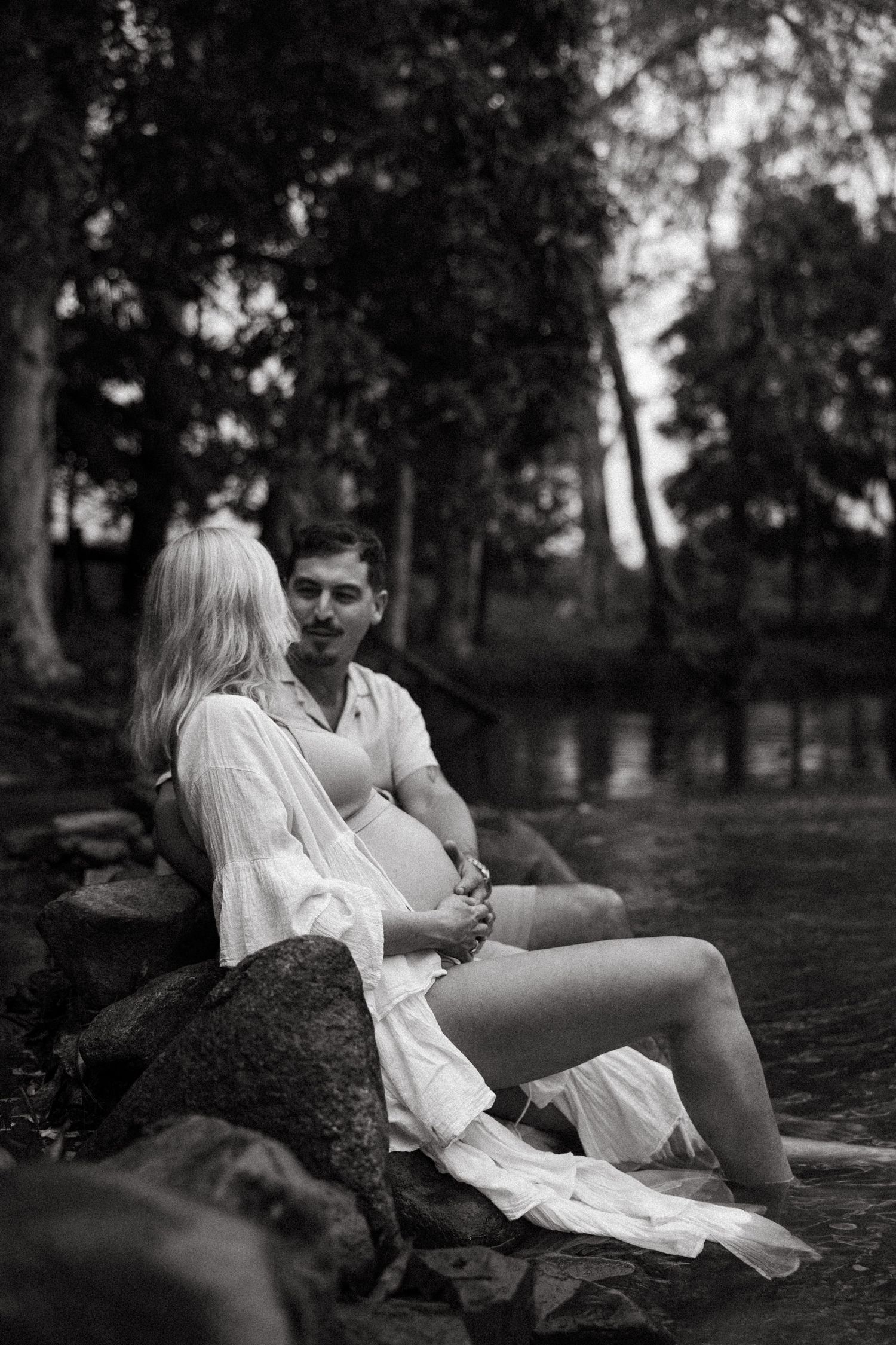 A romantic black and white photo of a couple sitting by a lake surrounded by trees in a natural setting.