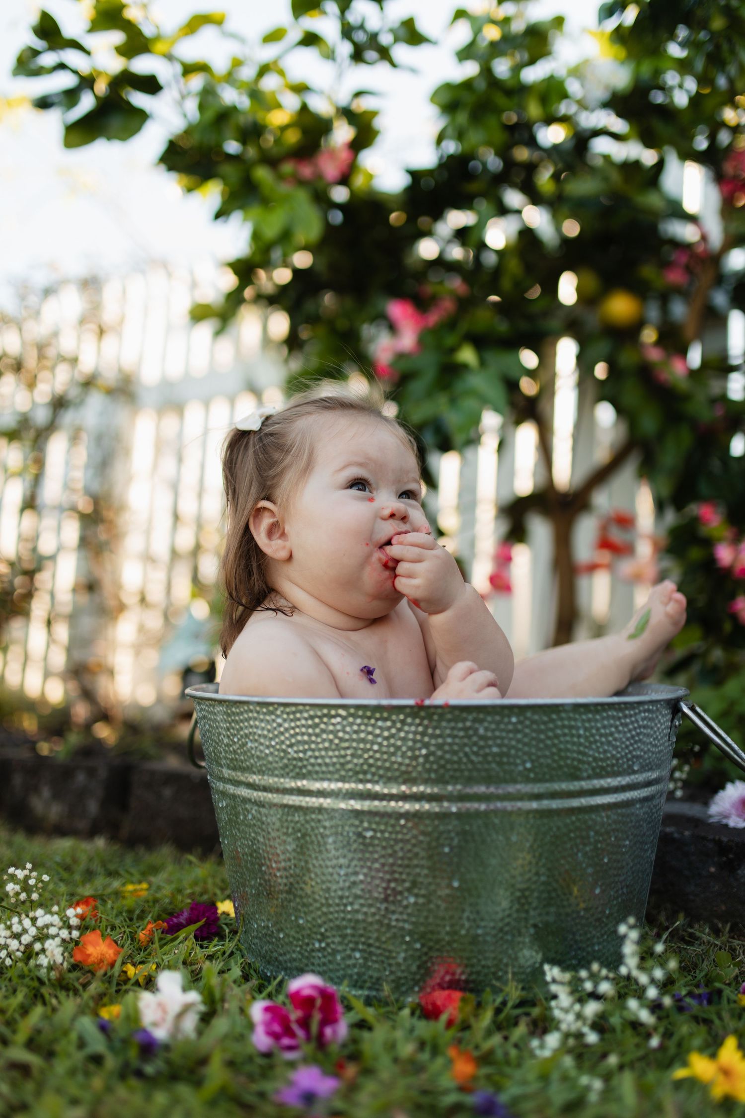 Baby playing in metal tub outdoors with flowers blooming in garden setting.