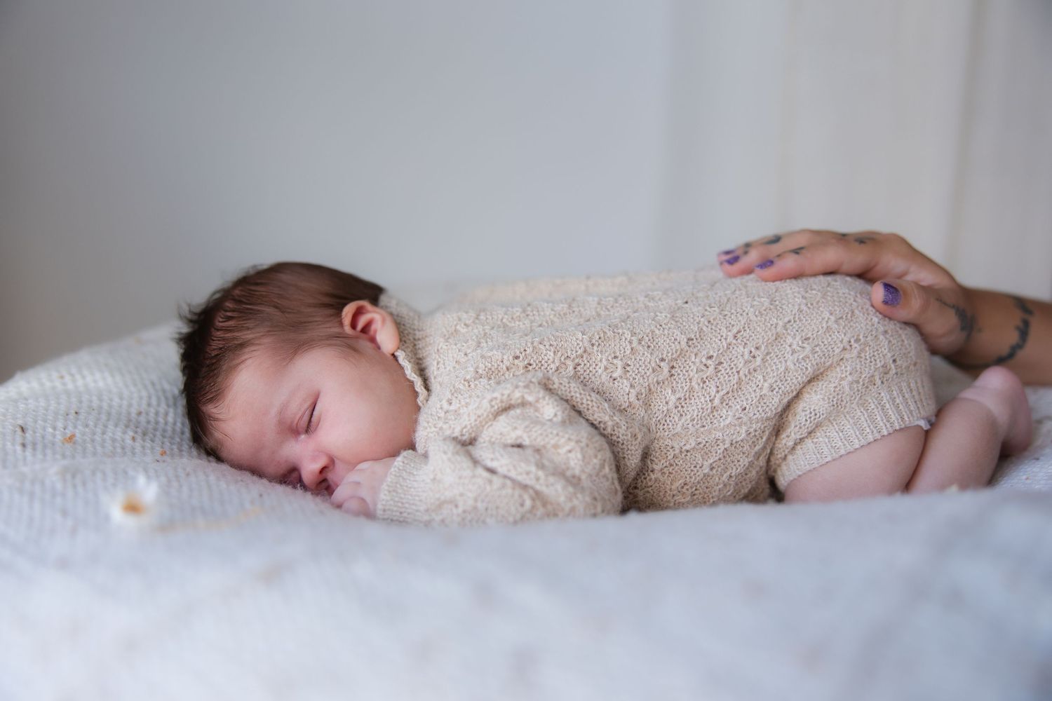 Sleeping newborn in cream knit outfit on white bedding with gentle hand touch.
