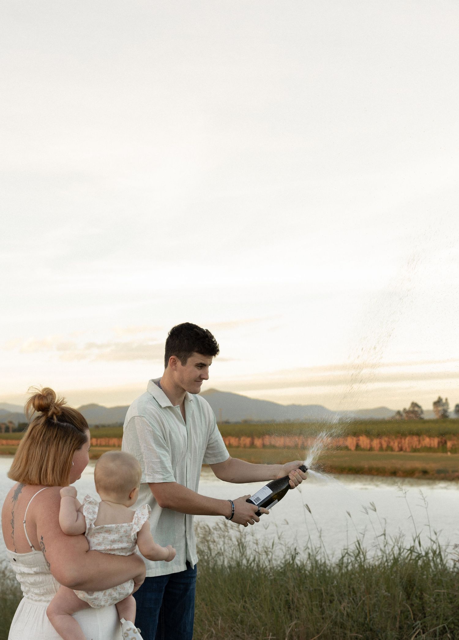 A family moment captured by a lake with someone opening a bottle during sunset.