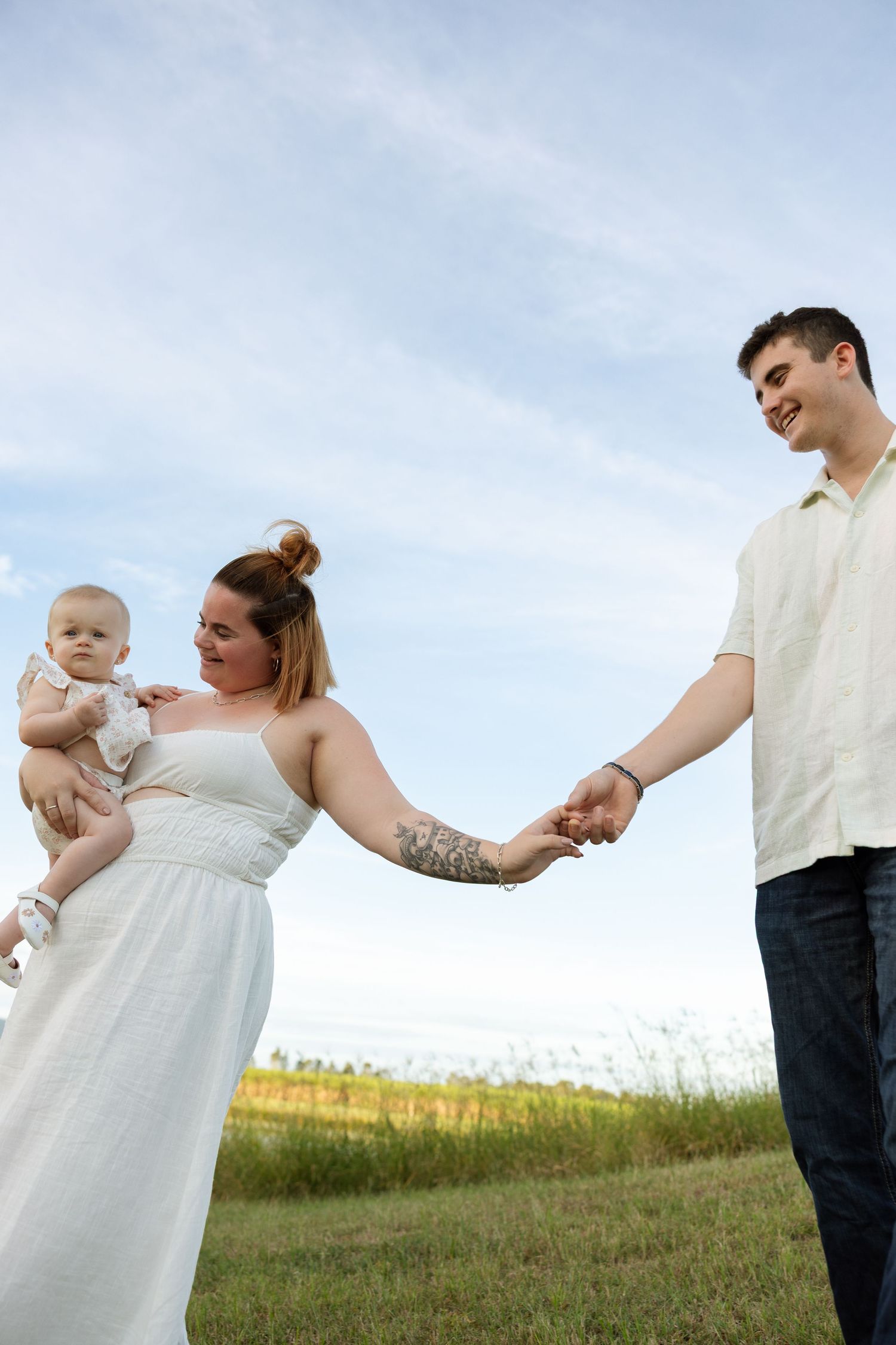 Young couple in white clothing holds hands while standing in a grassy field with their baby.
