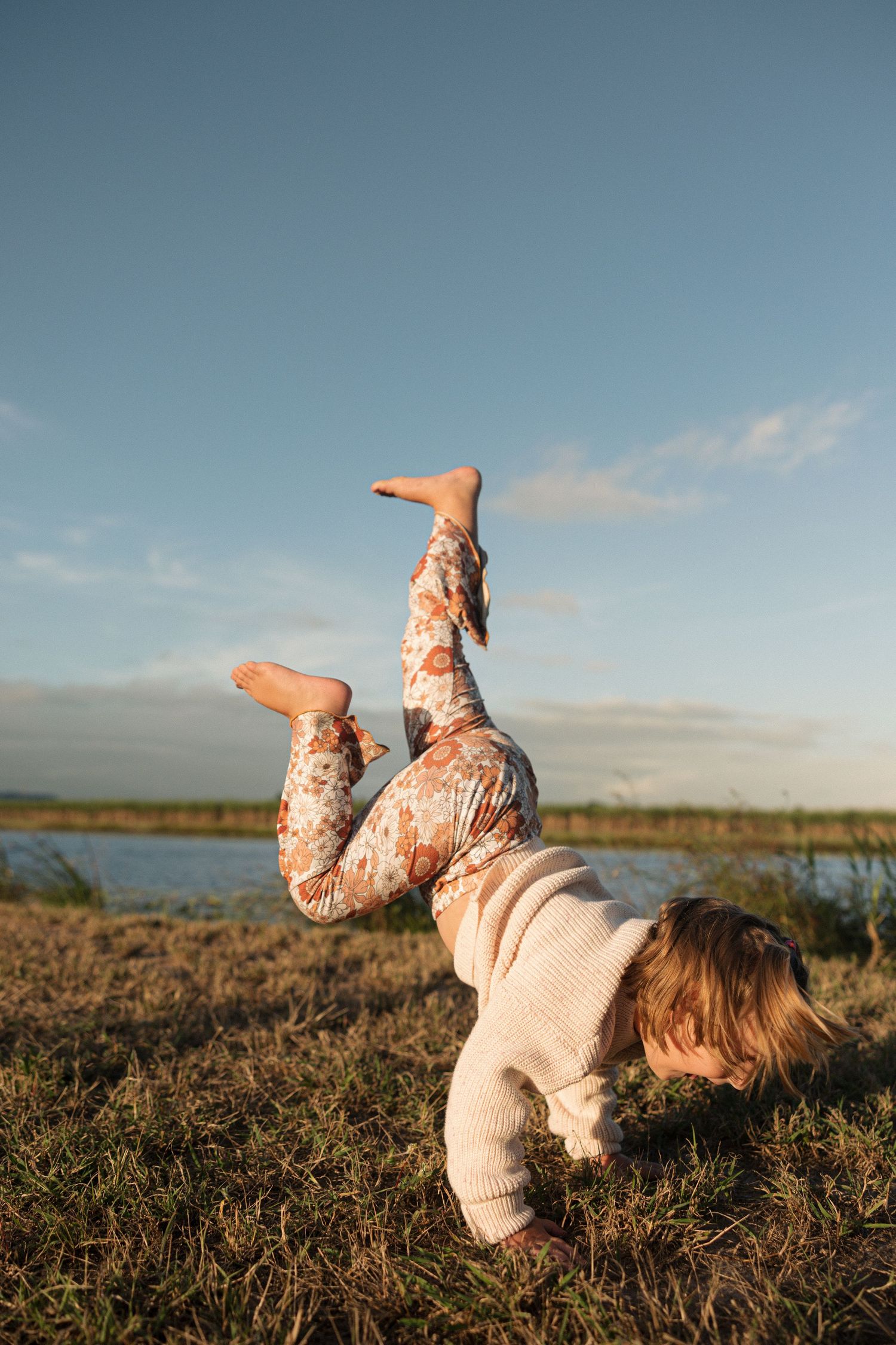 Playful handstand pose showcasing floral pants and beige sweater against a scenic waterfront backdrop.