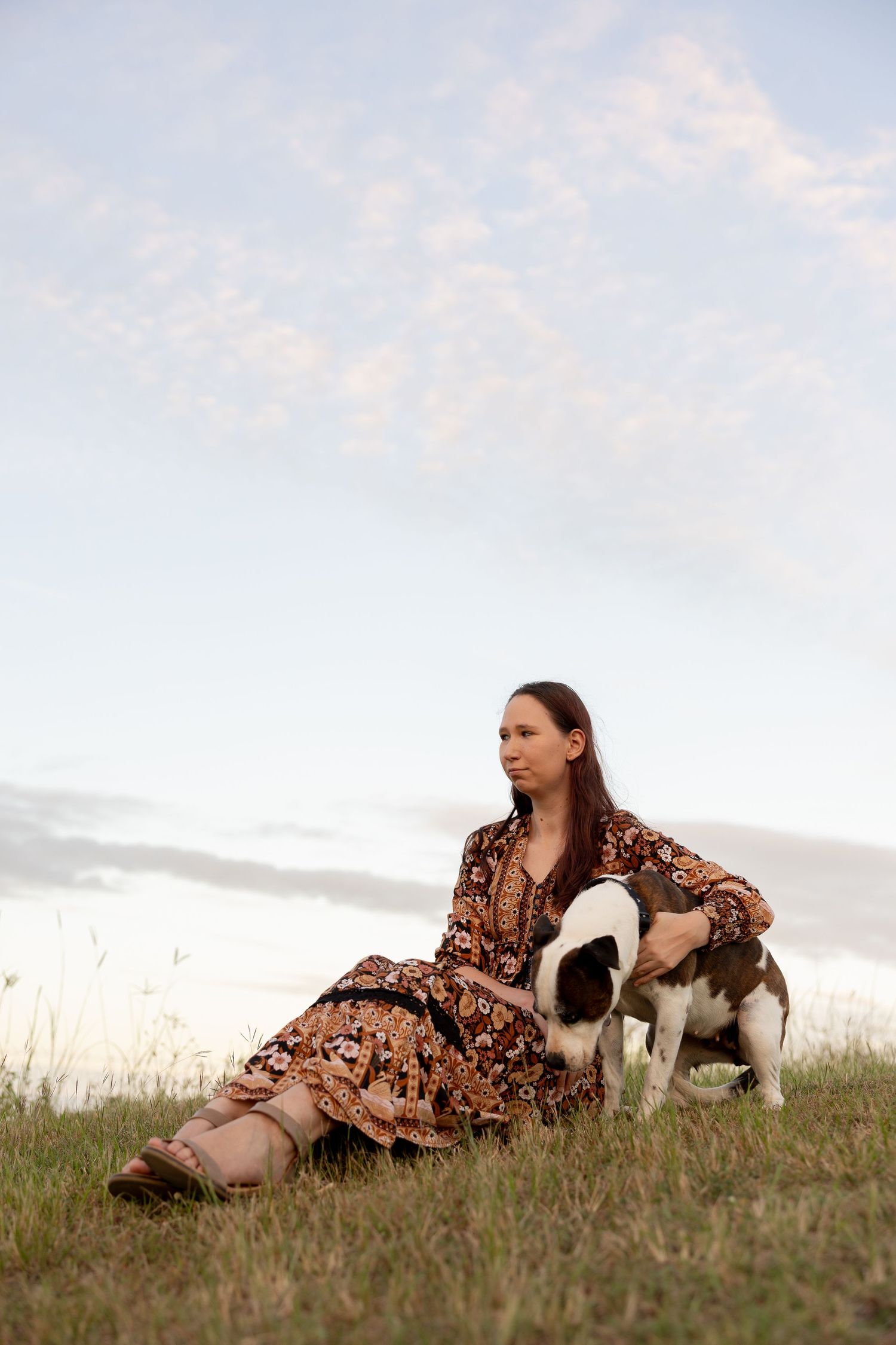 Person in floral dress rests in grassy field with black and white dog during sunset.