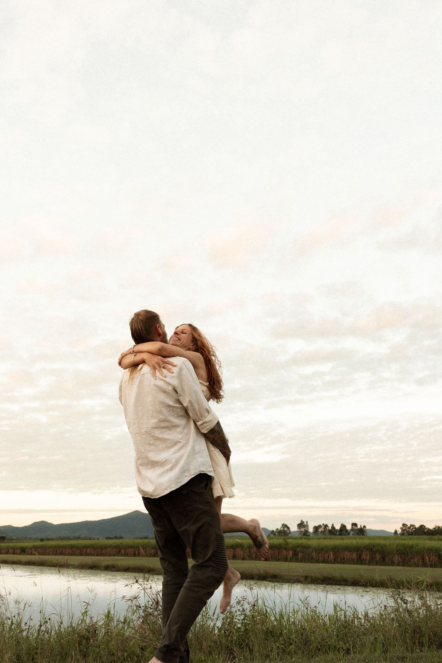 A couple embraces in a joyful hug against a scenic backdrop of mountains and a lake during sunset.