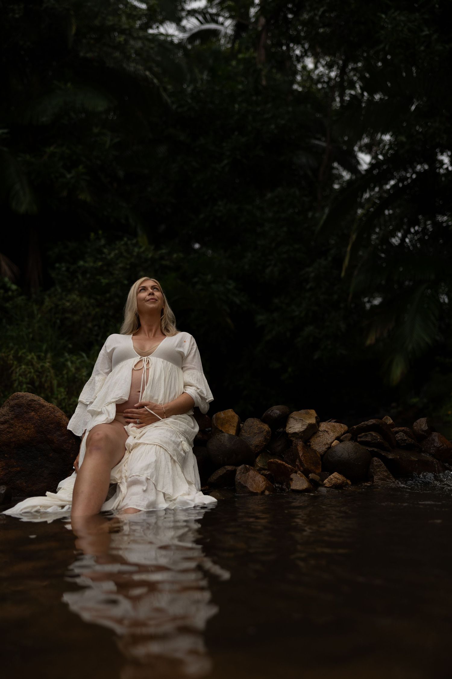Person in white dress sitting on logs by dark water in atmospheric forest setting.