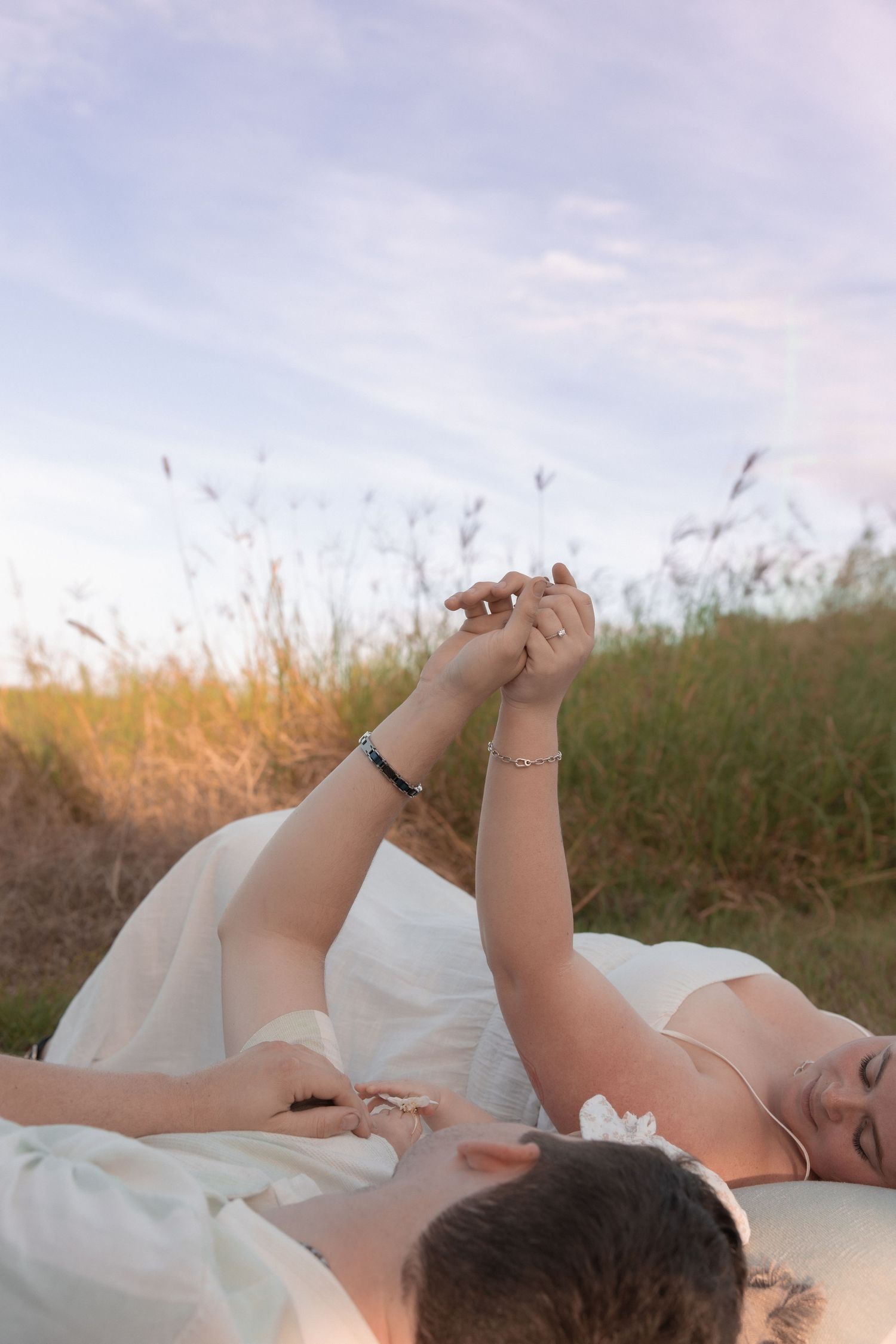 Couple lying in grass at sunset holding hands against soft sky.