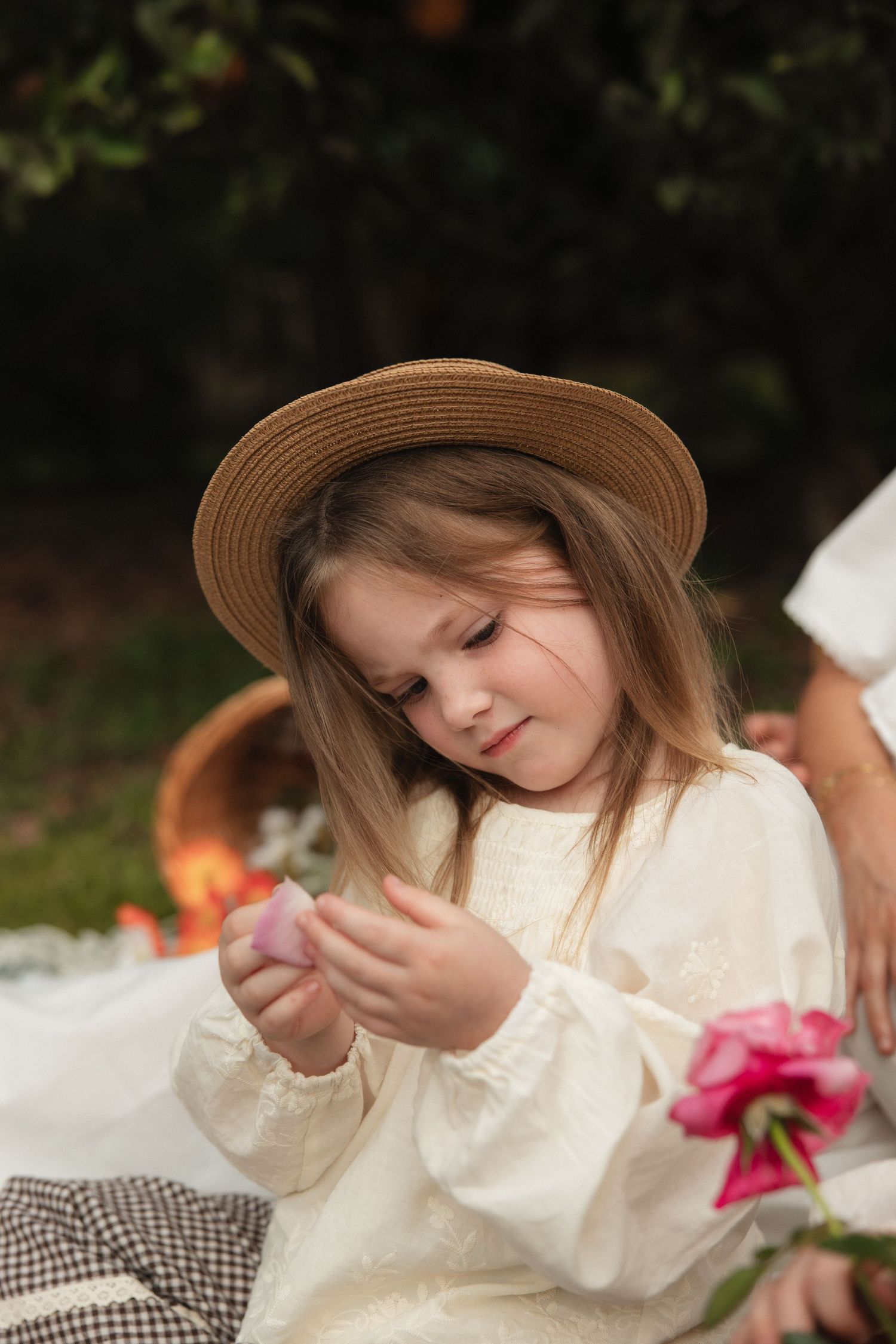 Child wearing a straw hat and cream sweater sits on a picnic blanket examining something small in their hands.