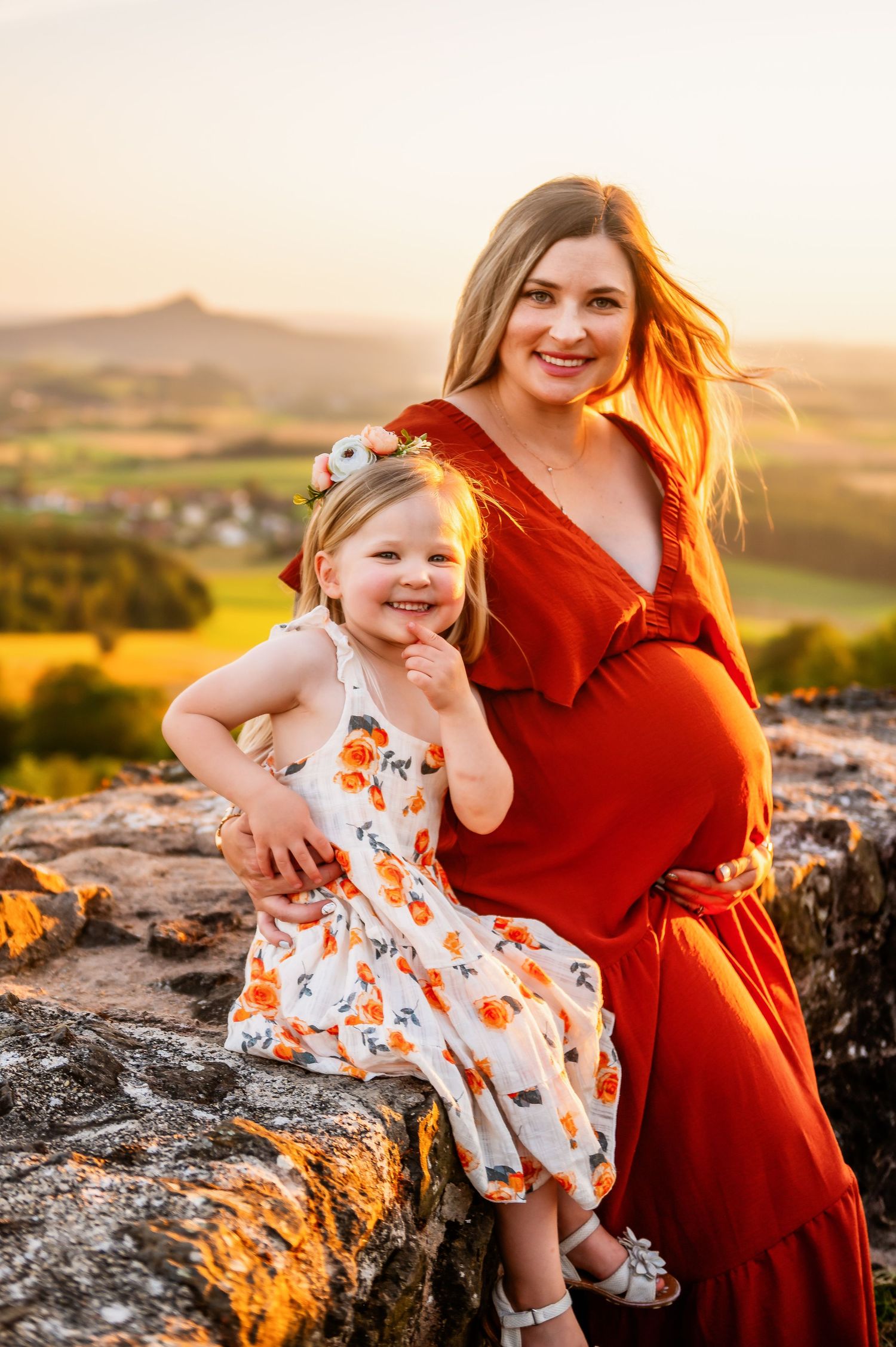 Mother and daughter share joyful moment on rocky overlook during sunset maternity session.