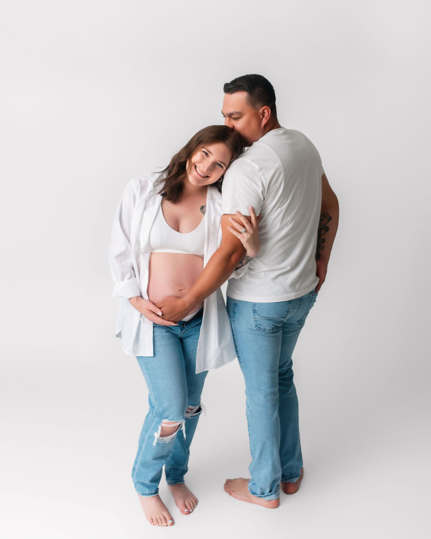 Barefoot couple in matching white tops and jeans pose for maternity photo.
