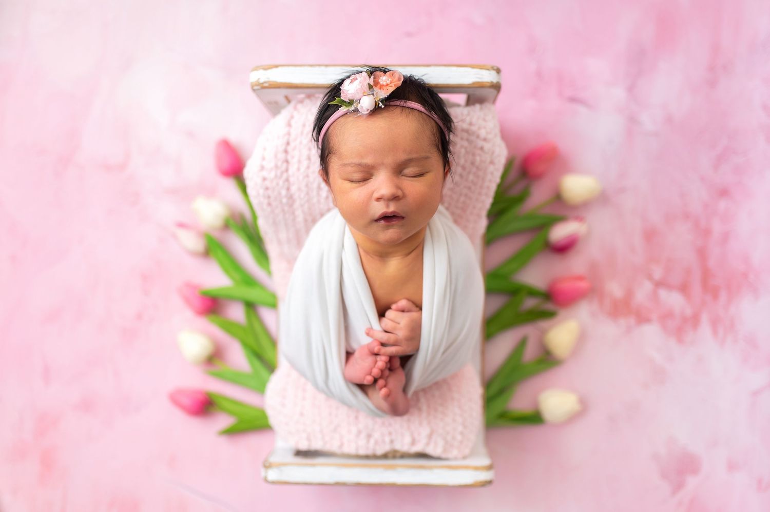 Sleeping newborn wrapped in white fabric lying on pink box with tulips in soft pink background.
