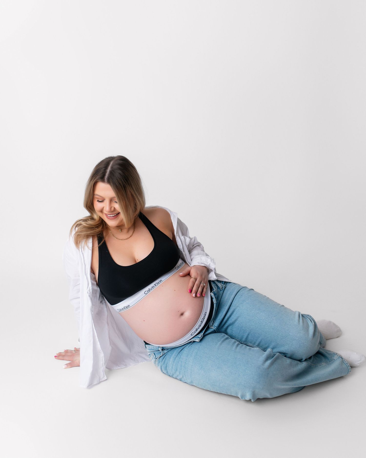 Expectant woman in black top and white cardigan poses on white background showing her pregnant belly.