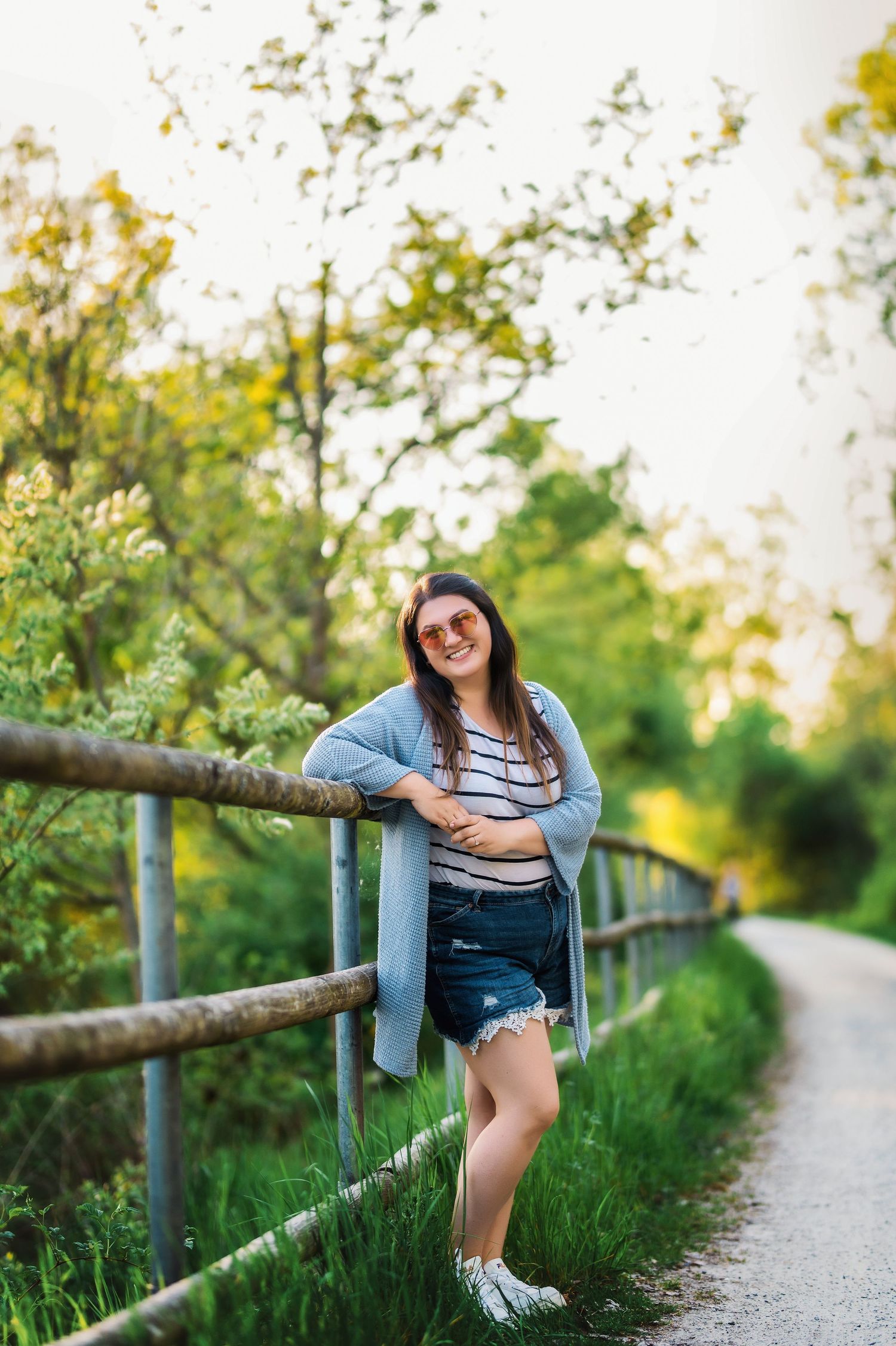 Casual portrait of person in denim cardigan posing by wooden railing on countryside path.