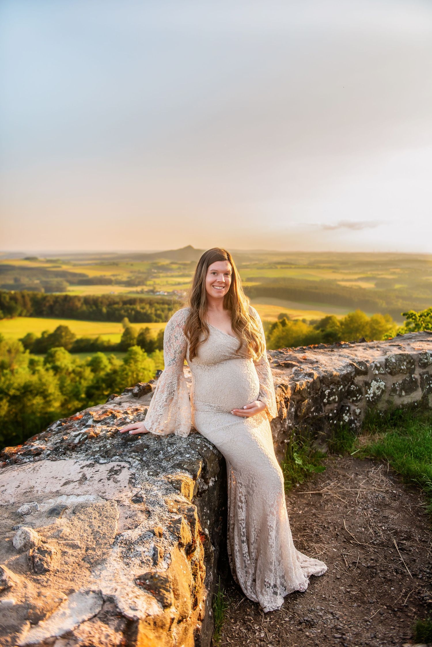 Pregnant woman in lace dress poses on rocky overlook during golden hour.