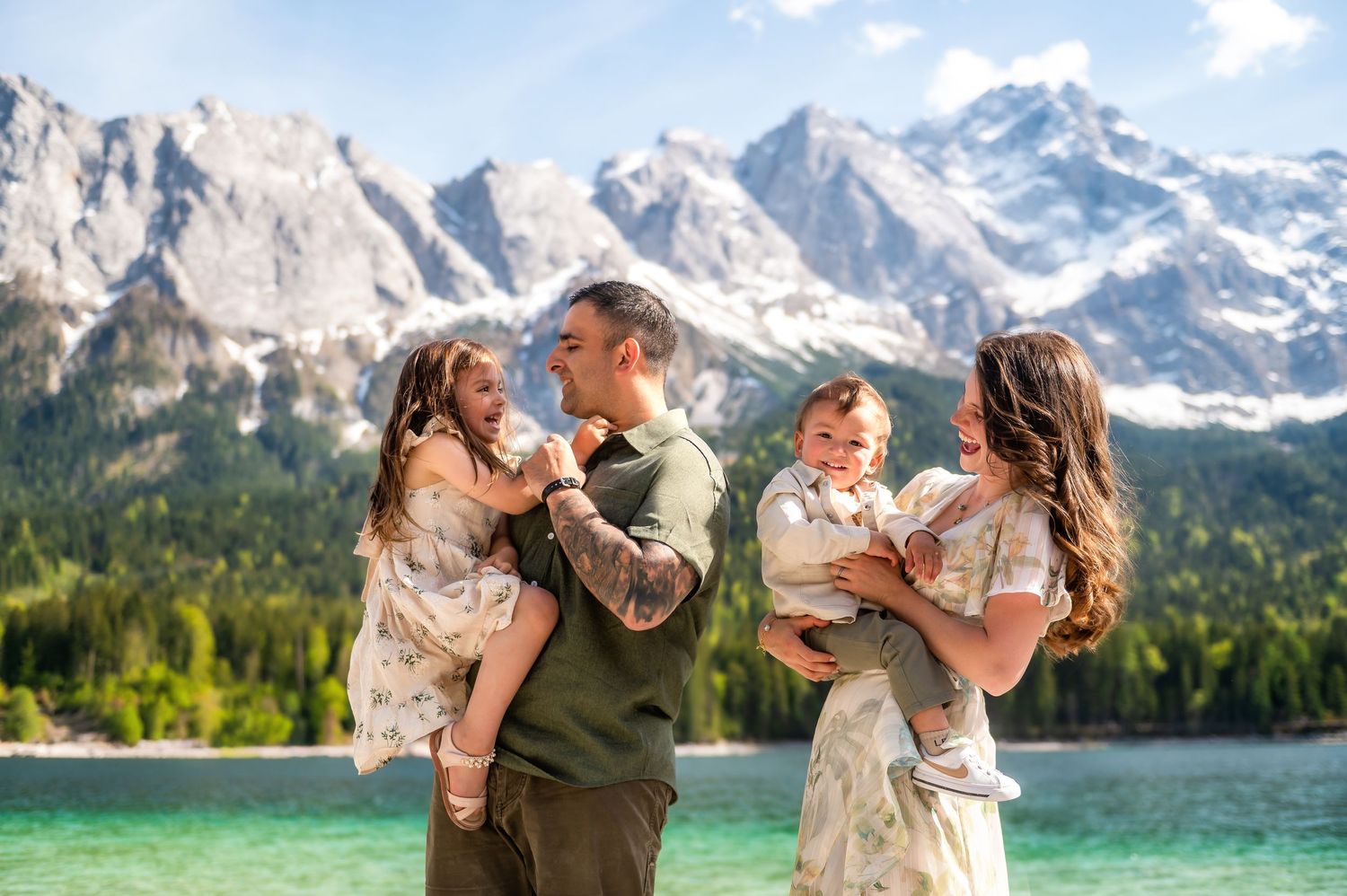 Family of four poses by a turquoise alpine lake with snow-capped Dolomite mountains in the background.