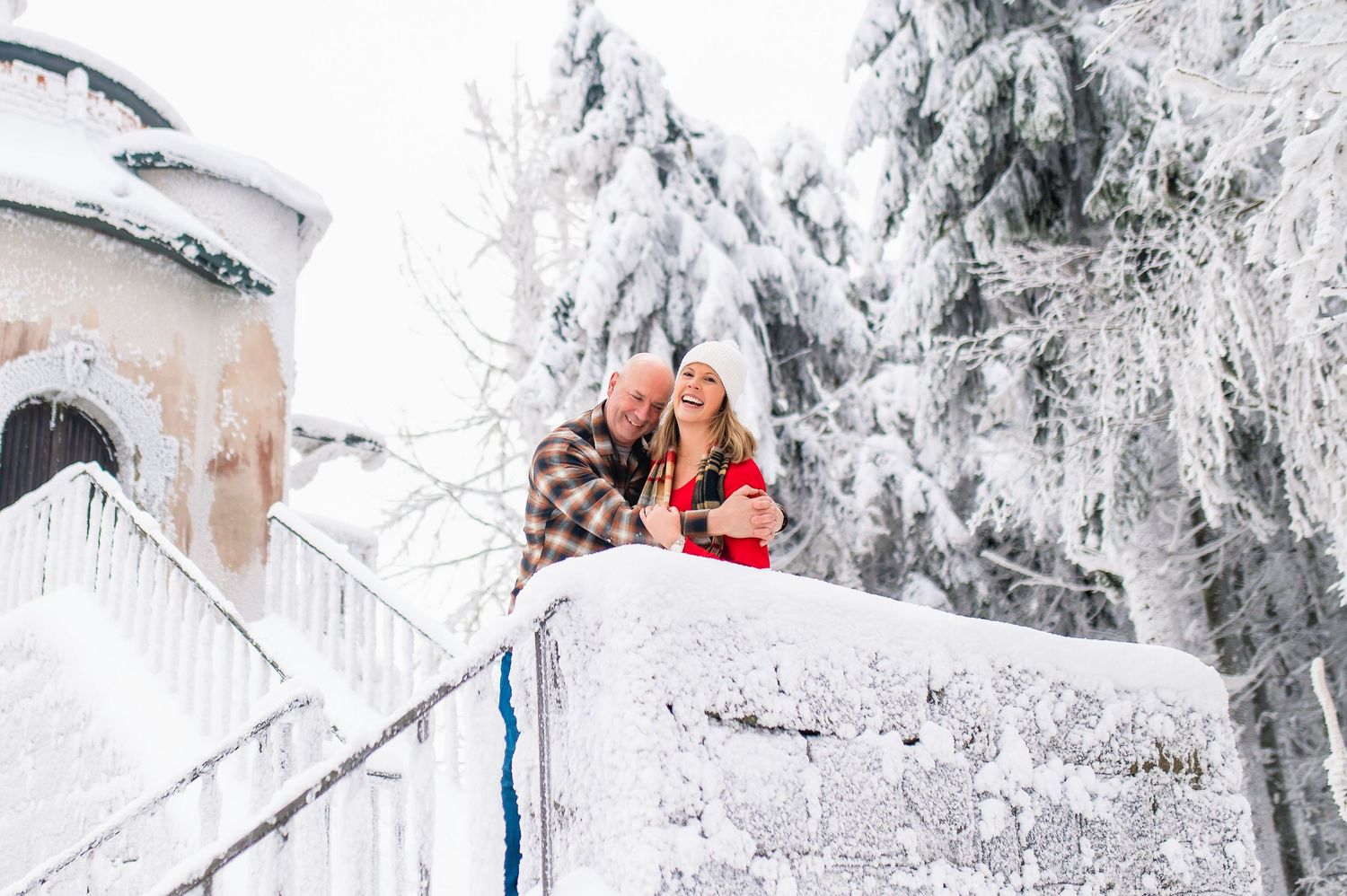 Two people and a dog sit on snowy steps surrounded by frost-covered trees in a winter wonderland setting.