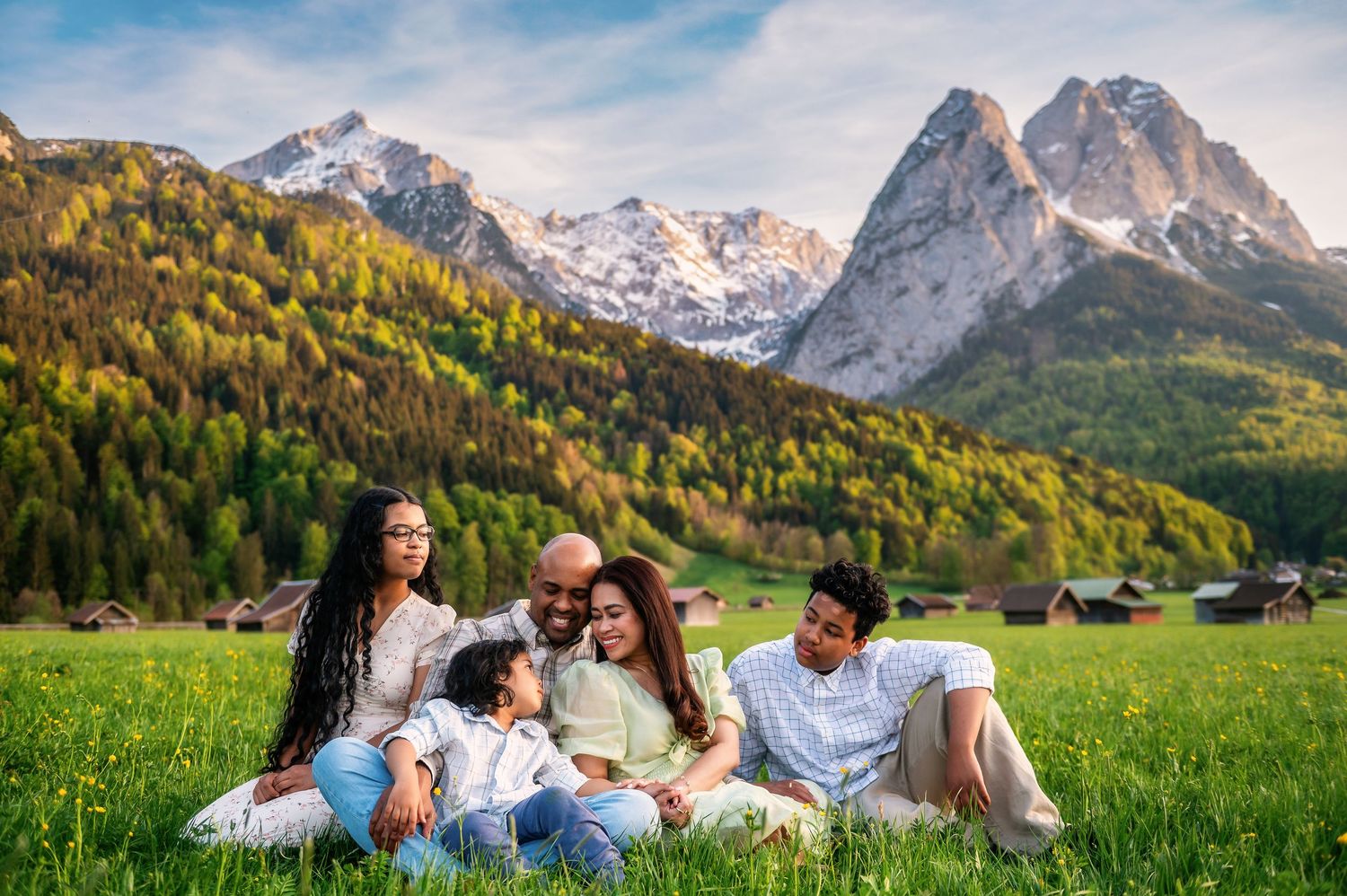 Happy multiracial family sitting together on green meadow with snow-capped Alpine mountains and forest in background.