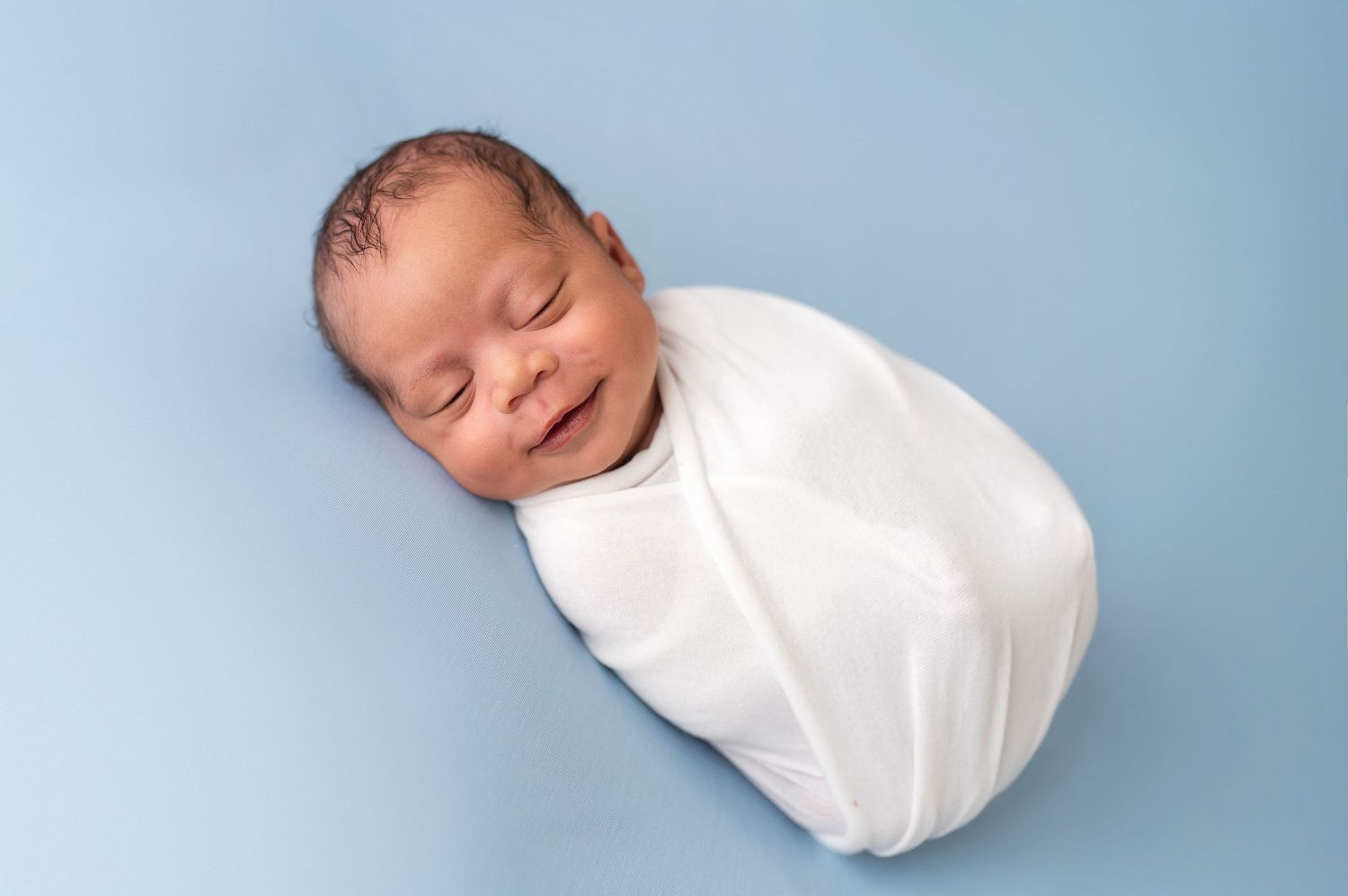 Peaceful sleeping newborn swaddled in white wrap against blue background.