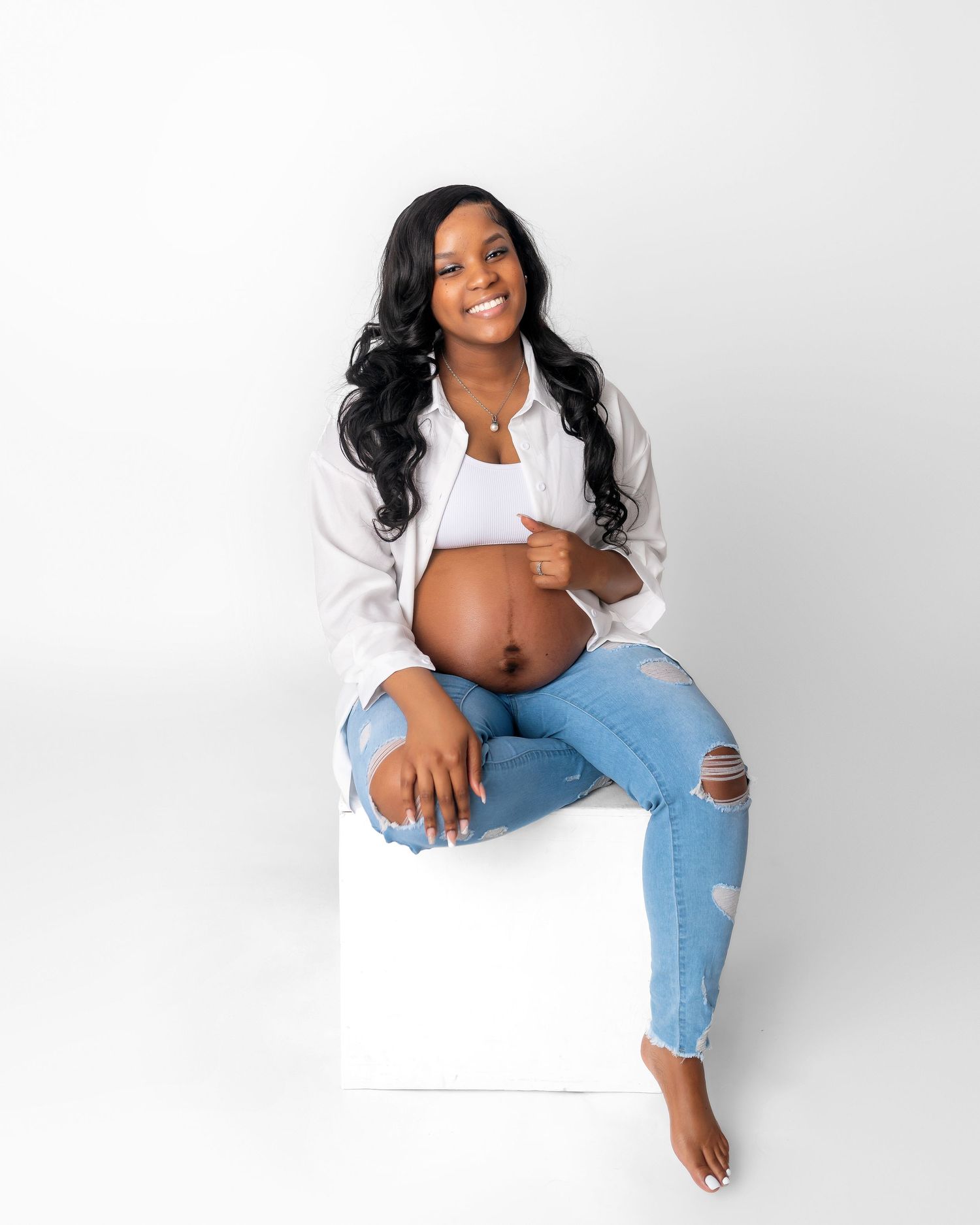 Pregnant woman in white shirt and ripped jeans sits casually on white cube during maternity photoshoot.