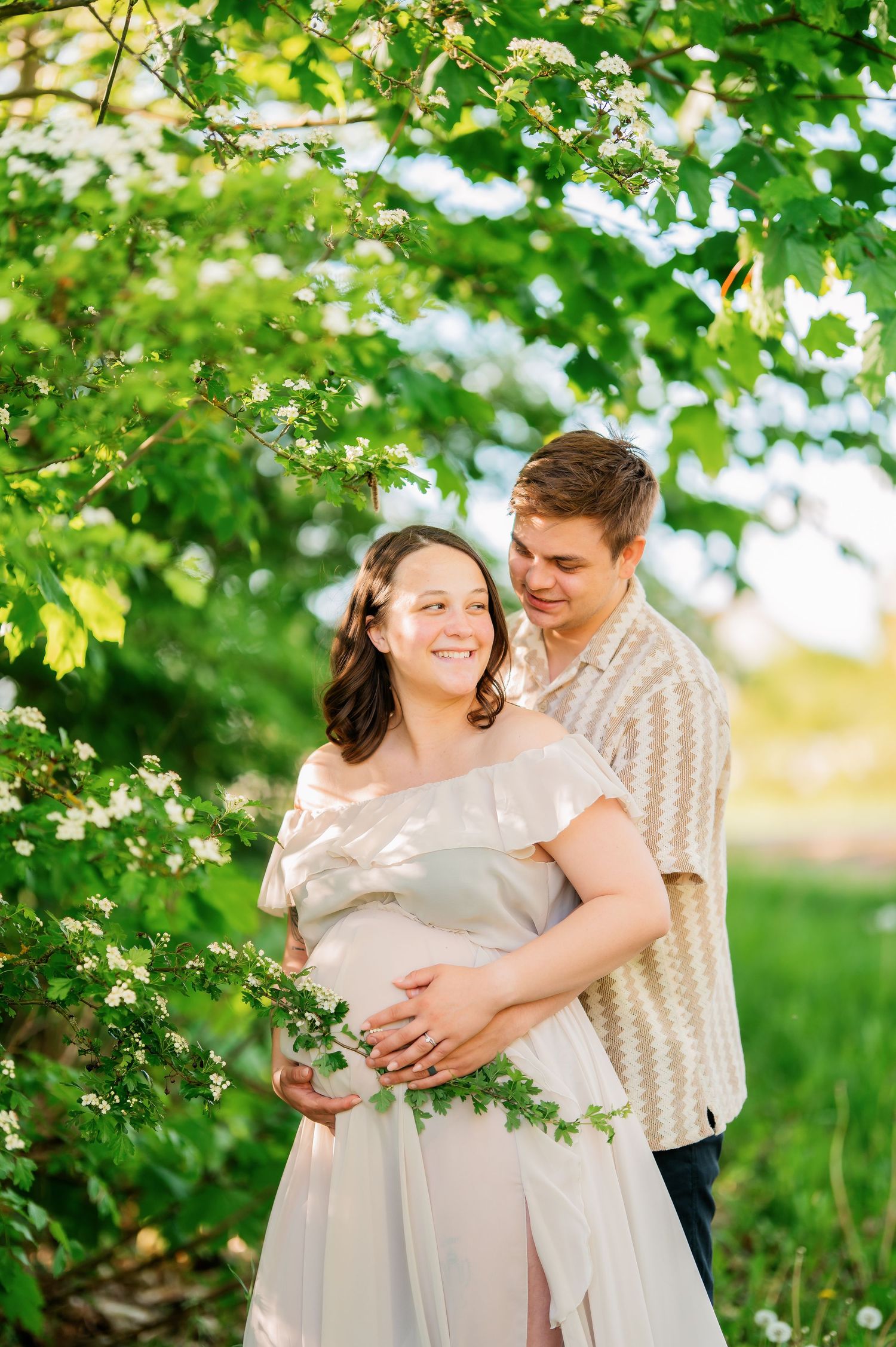Romantic spring maternity photos with couple embracing under green leafy trees.