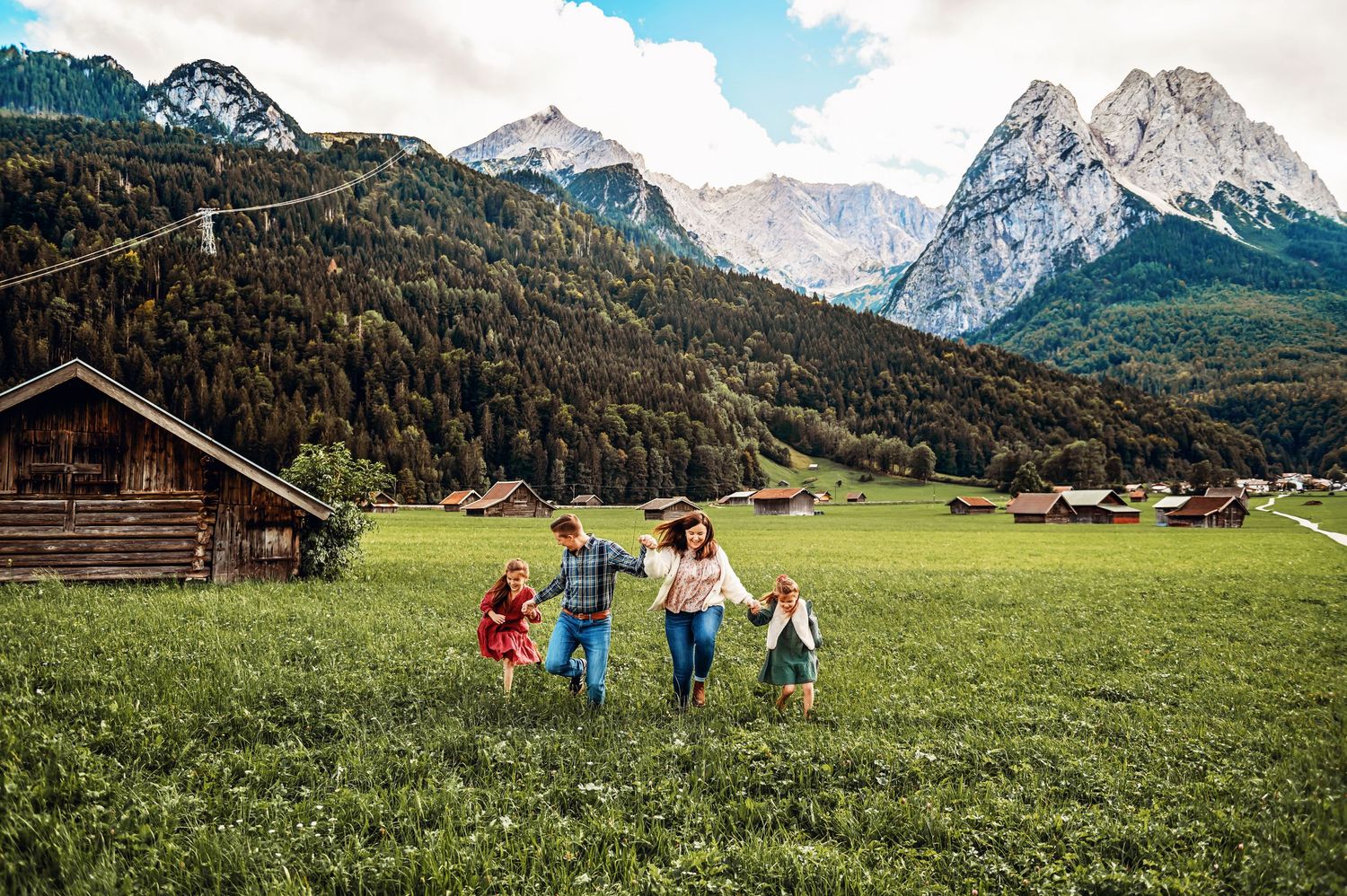 Family walking through green alpine meadow with rustic wooden barns and dramatic snow-capped mountain peaks in background.