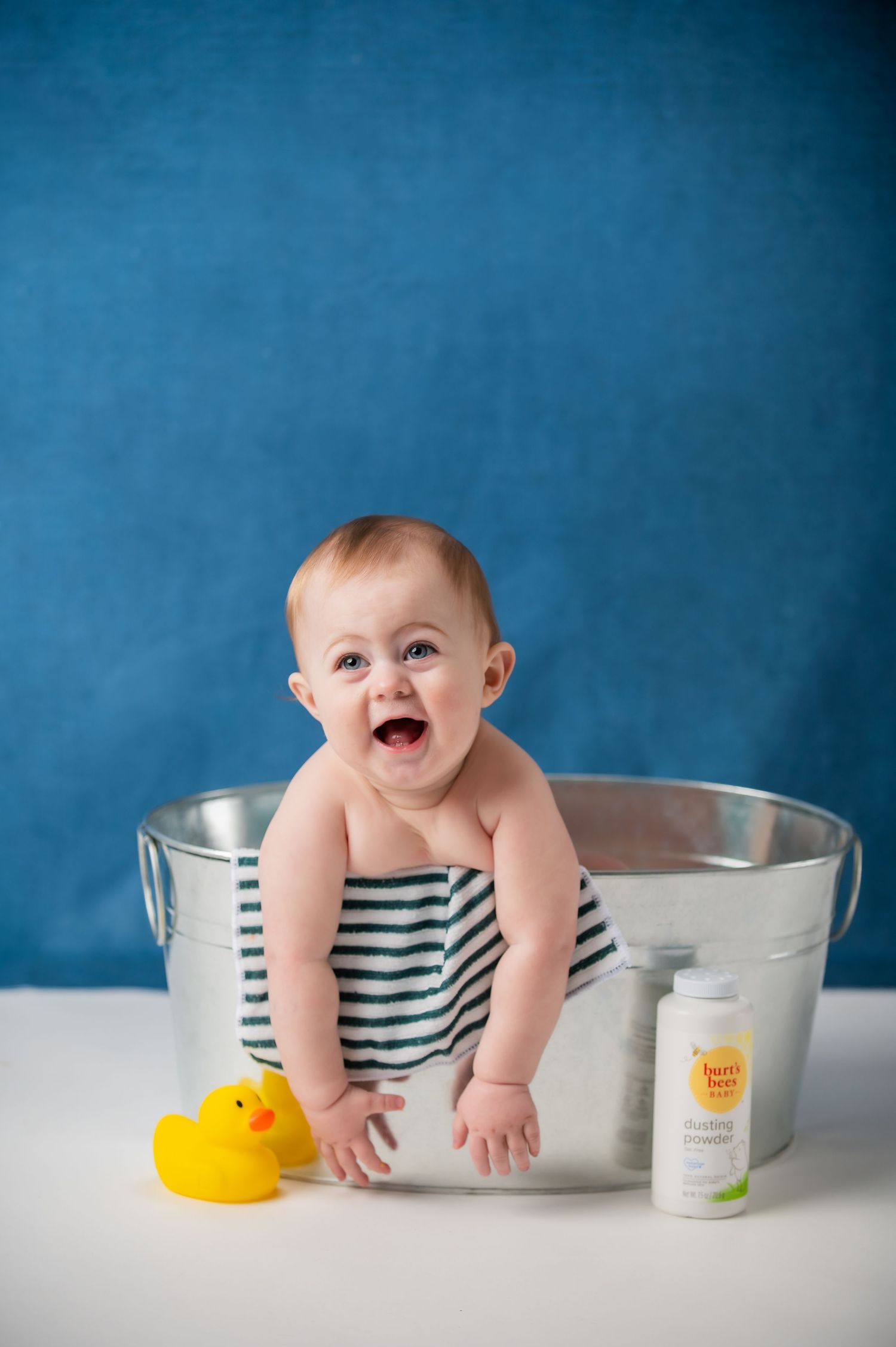 Happy baby sitting in a metal tub with a rubber duck and baby shampoo on a blue background.