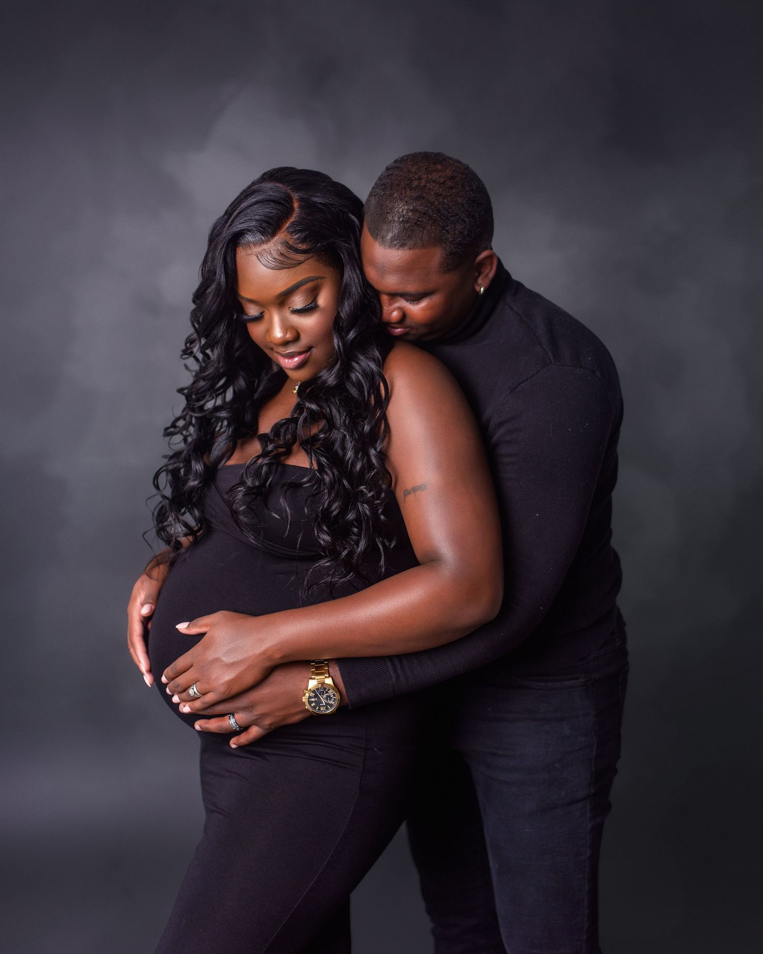 Intimate maternity portrait of couple in black attire embracing against dark background.