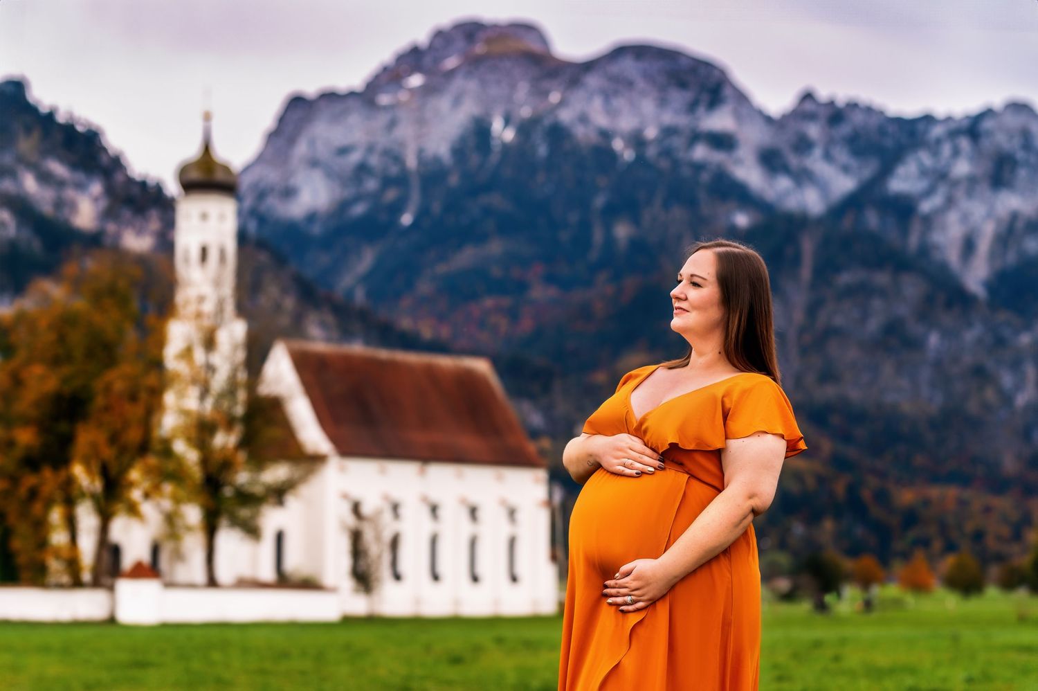 A pair embraces on a green meadow with a scenic mountain landscape and white church steeple in the distance.