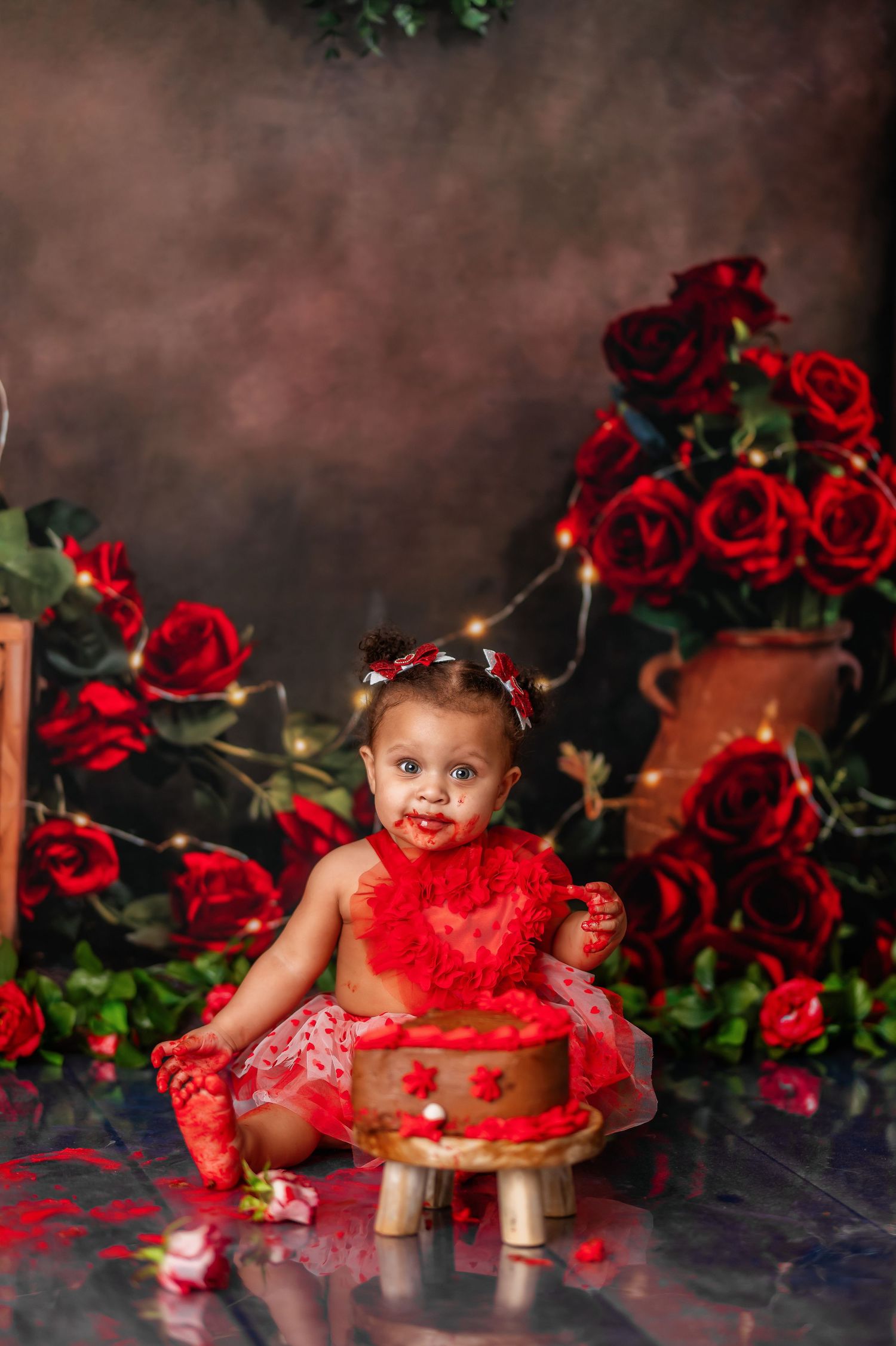 Baby in red dress crawling near cake during romantic Valentine's photoshoot with roses.
