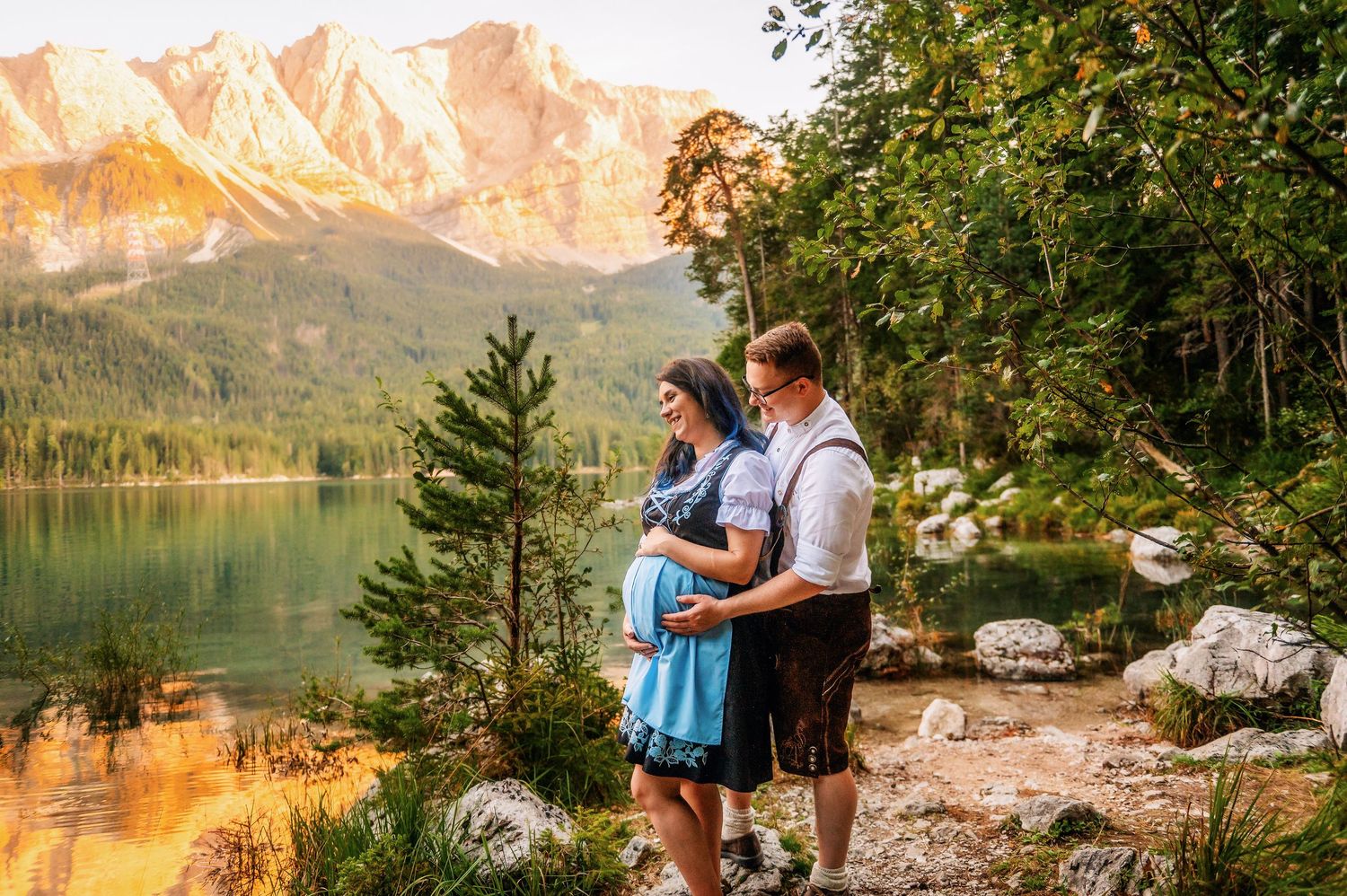 Romantic couple sitting on rocky shore by mountain lake during golden sunset.