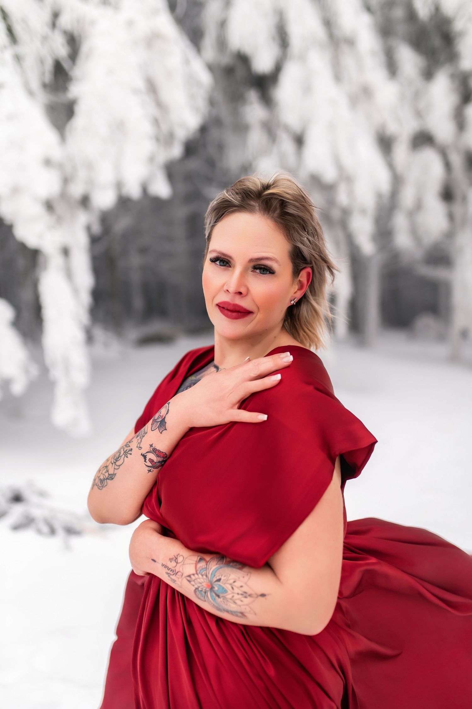 Woman in red dress with tattoos poses in snowy winter forest, wearing red lipstick and elegant updo hairstyle.