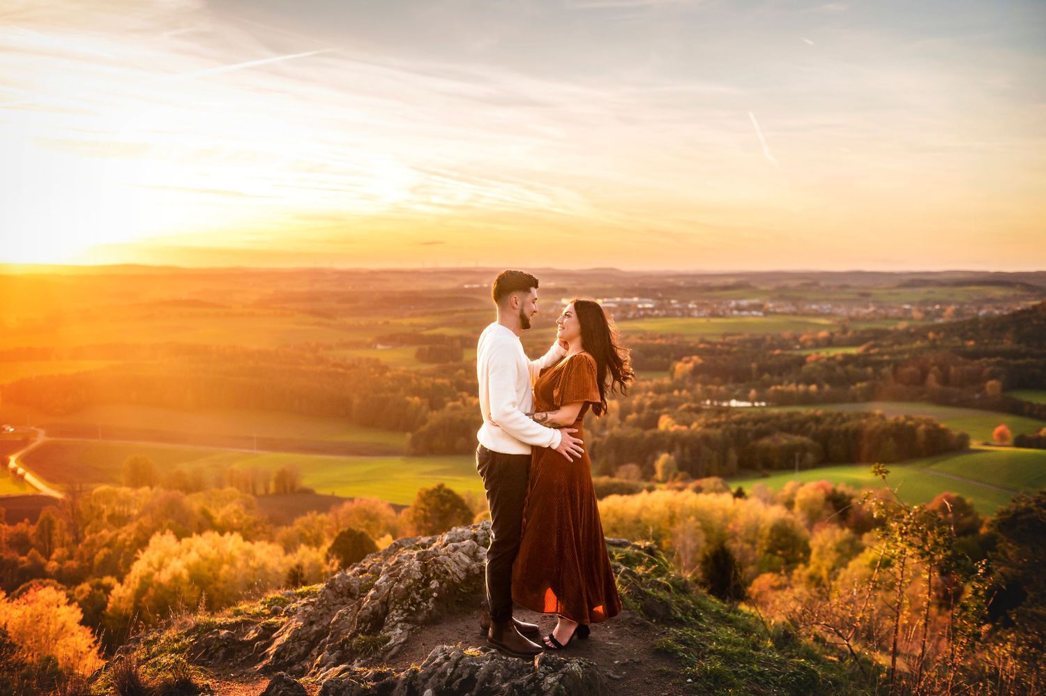 Couple embracing on rocky hilltop at golden sunset with scenic countryside valley view below.