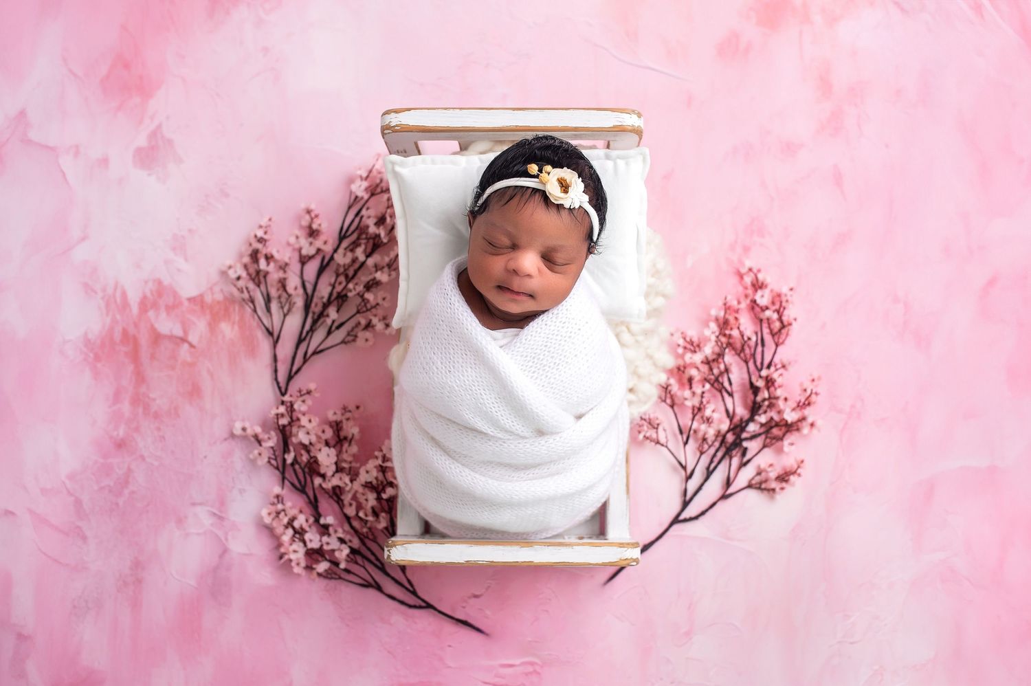 Newborn baby swaddled in white wrap sleeping in a tiny wooden bed with pink cherry blossoms on a pink background.