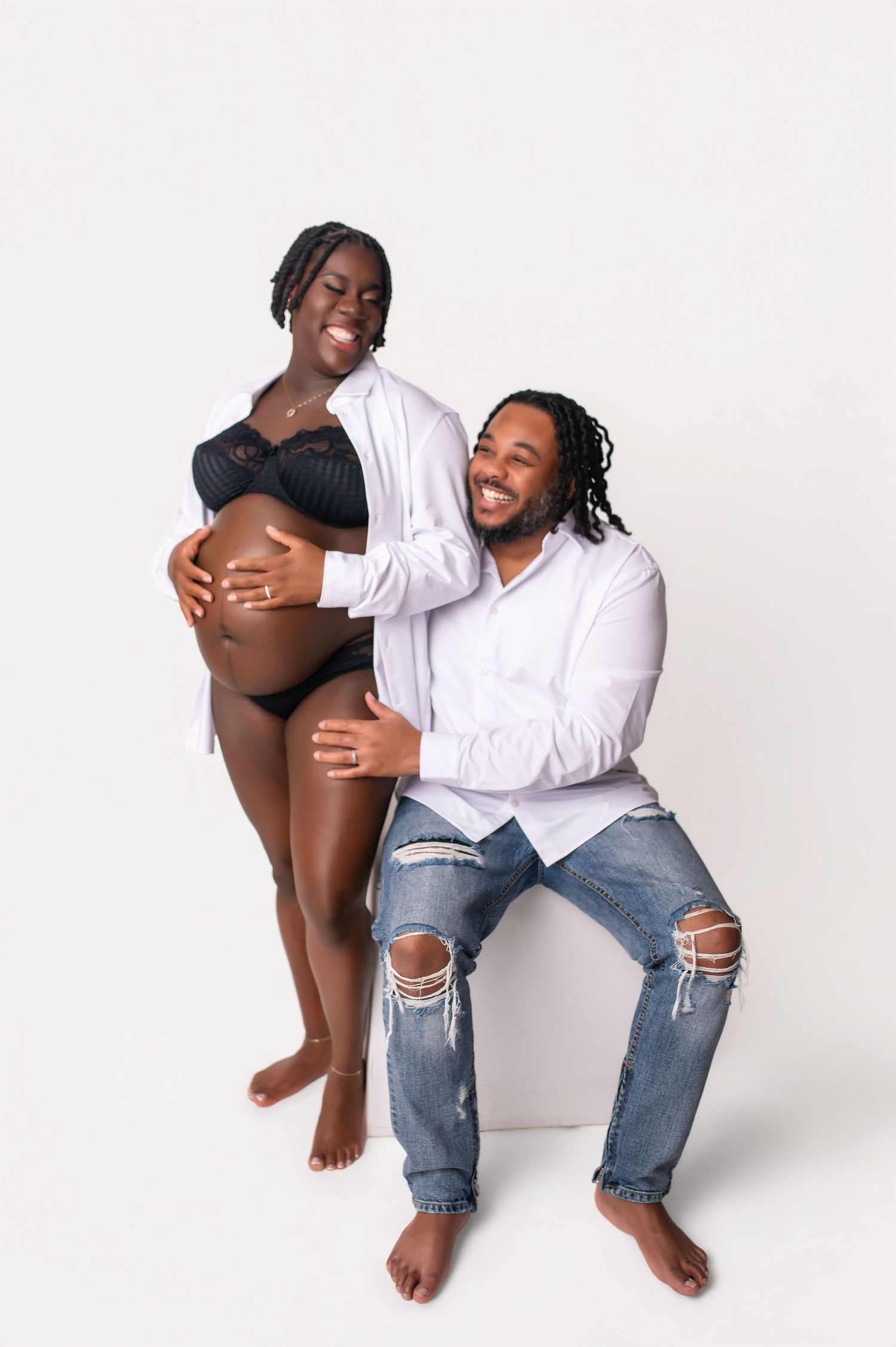 Happy couple expecting a baby, man in white shirt and jeans sits beside pregnant woman in black lingerie.