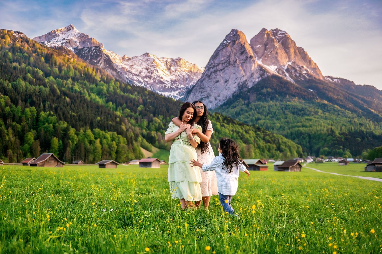 Family embracing in a lush green meadow with snow-capped mountains and pine forests in the background.